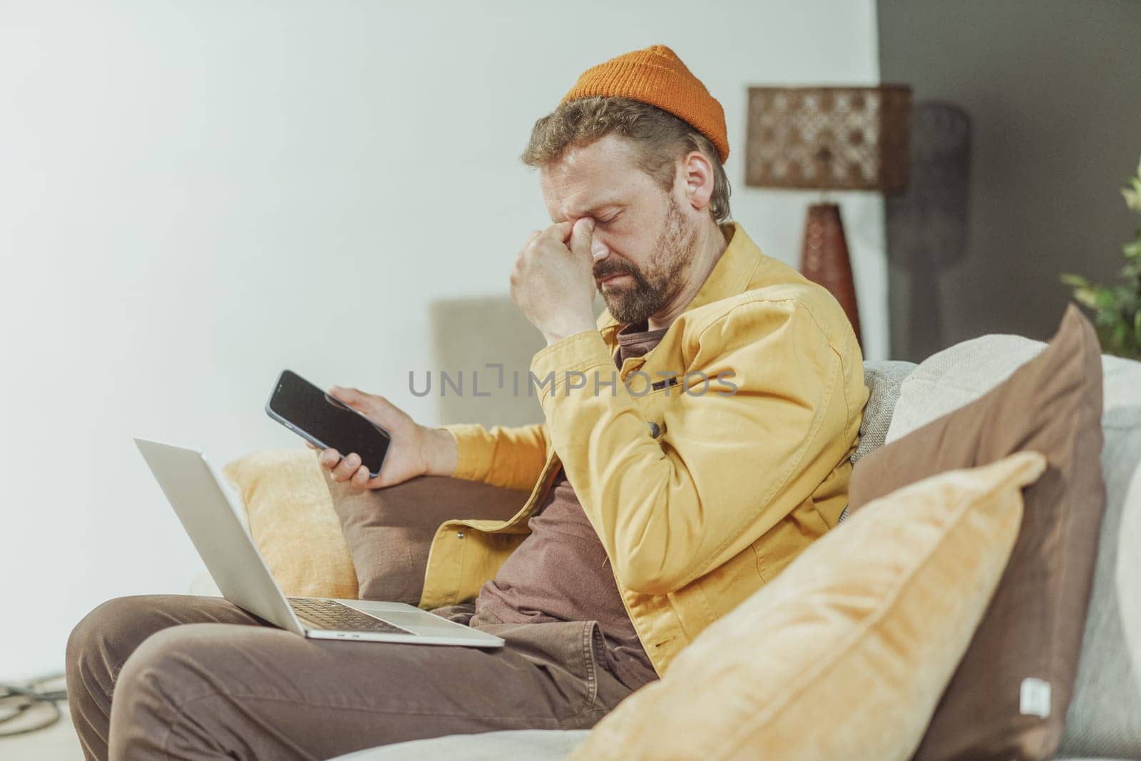 Tired man finds solace on his home sofa after session of browsing internet using both his mobile phone and laptop. Modern digital fatigue, as seeks rest and relaxation within comforts of his home. High quality photo