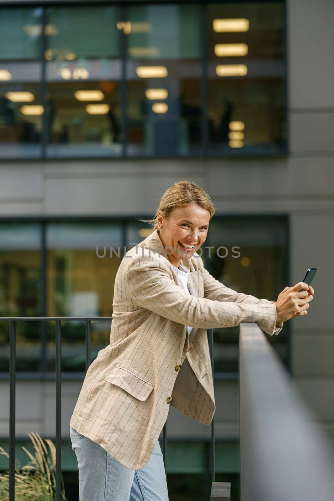 Smiling business woman holding phone while standing on modern office terrace and looks camera