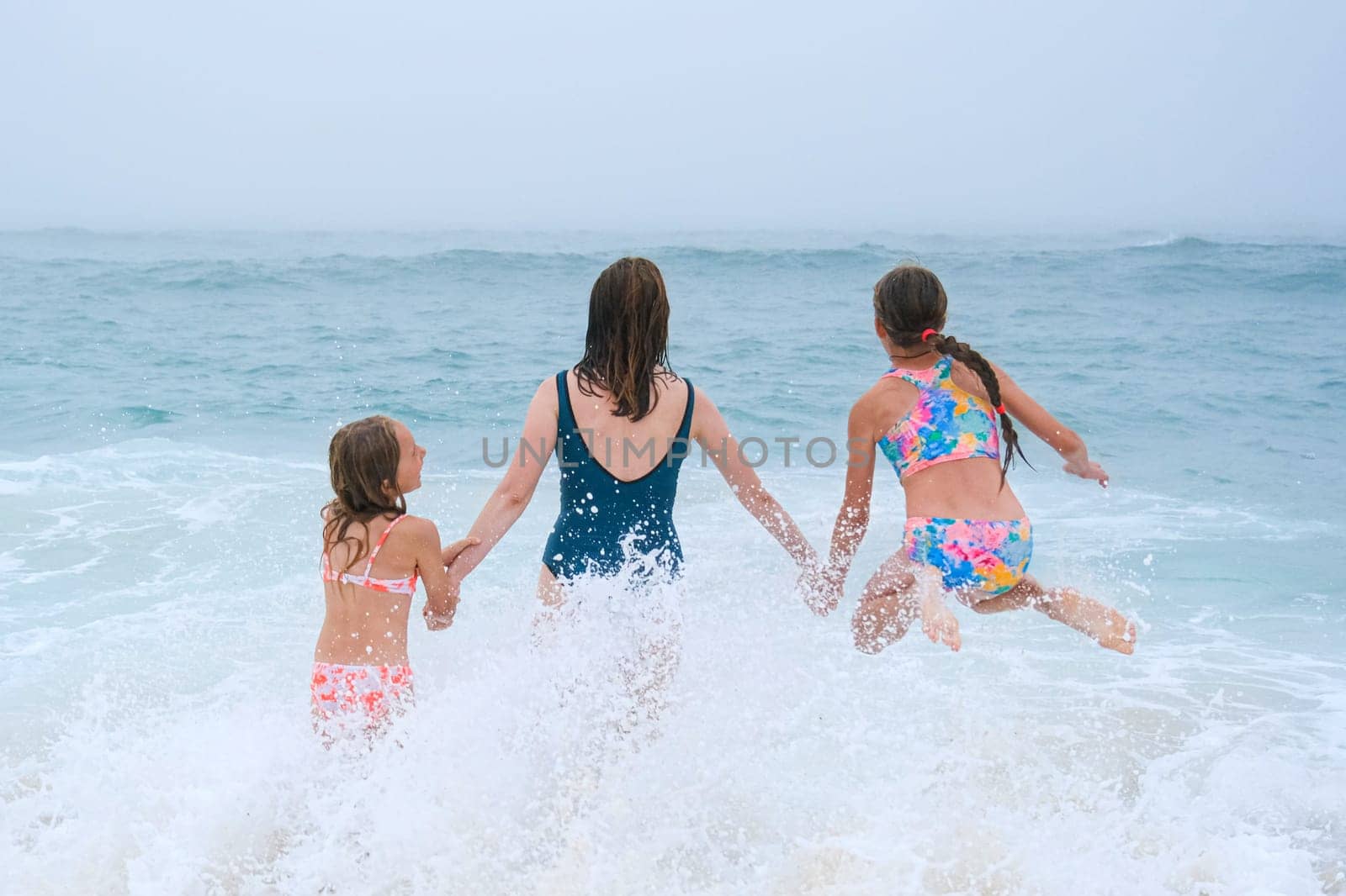 Mother and children playing on the ocean beach. Family enjoying the ocean. Mother holds girls's hands and they all look at the ocean together by esvetleishaya