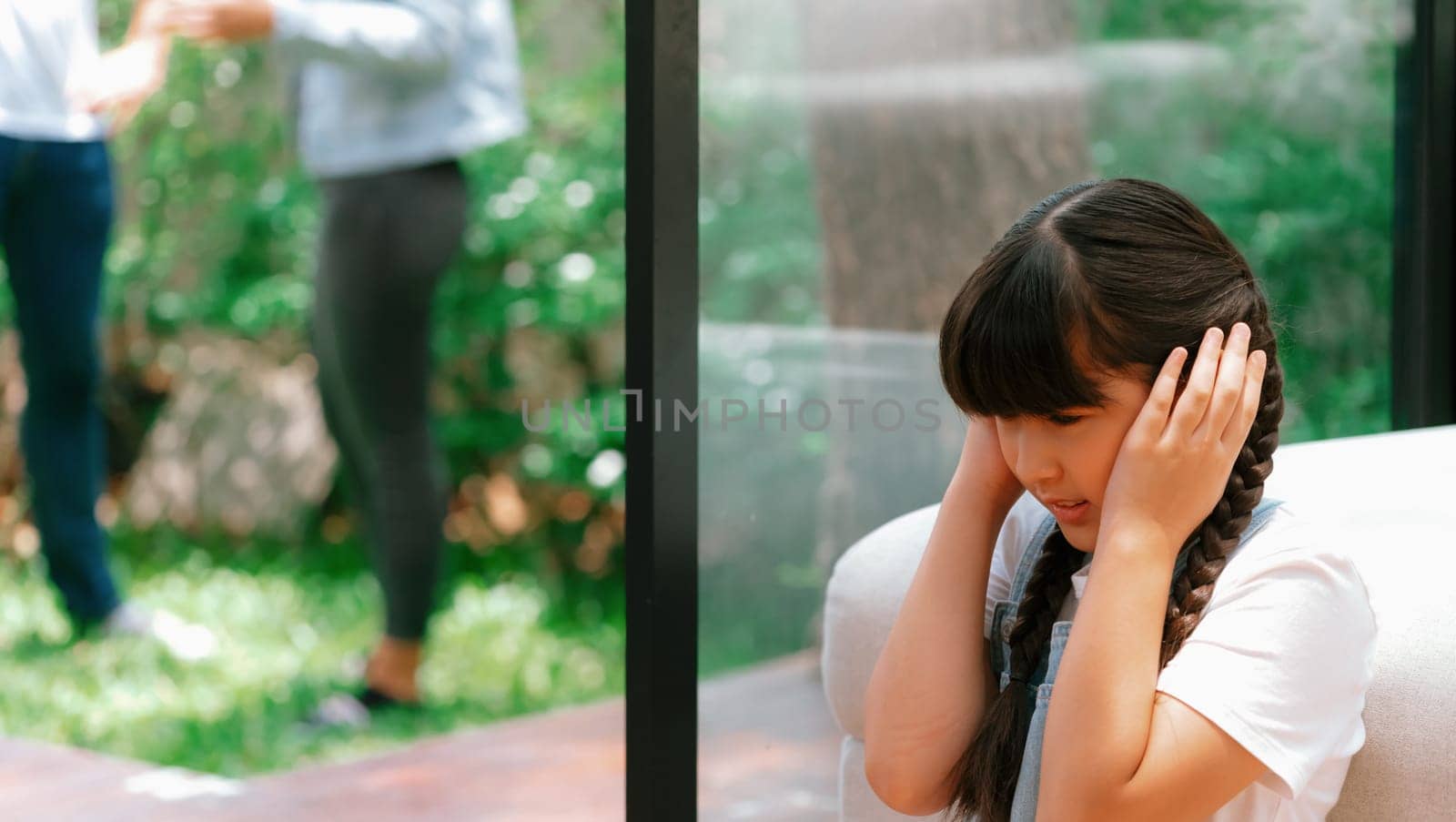 Stressed and unhappy young girl huddle in corner, cover her ears blocking sound of her parent arguing in background. Domestic violence at home and traumatic childhood develop to depression. Synchronos