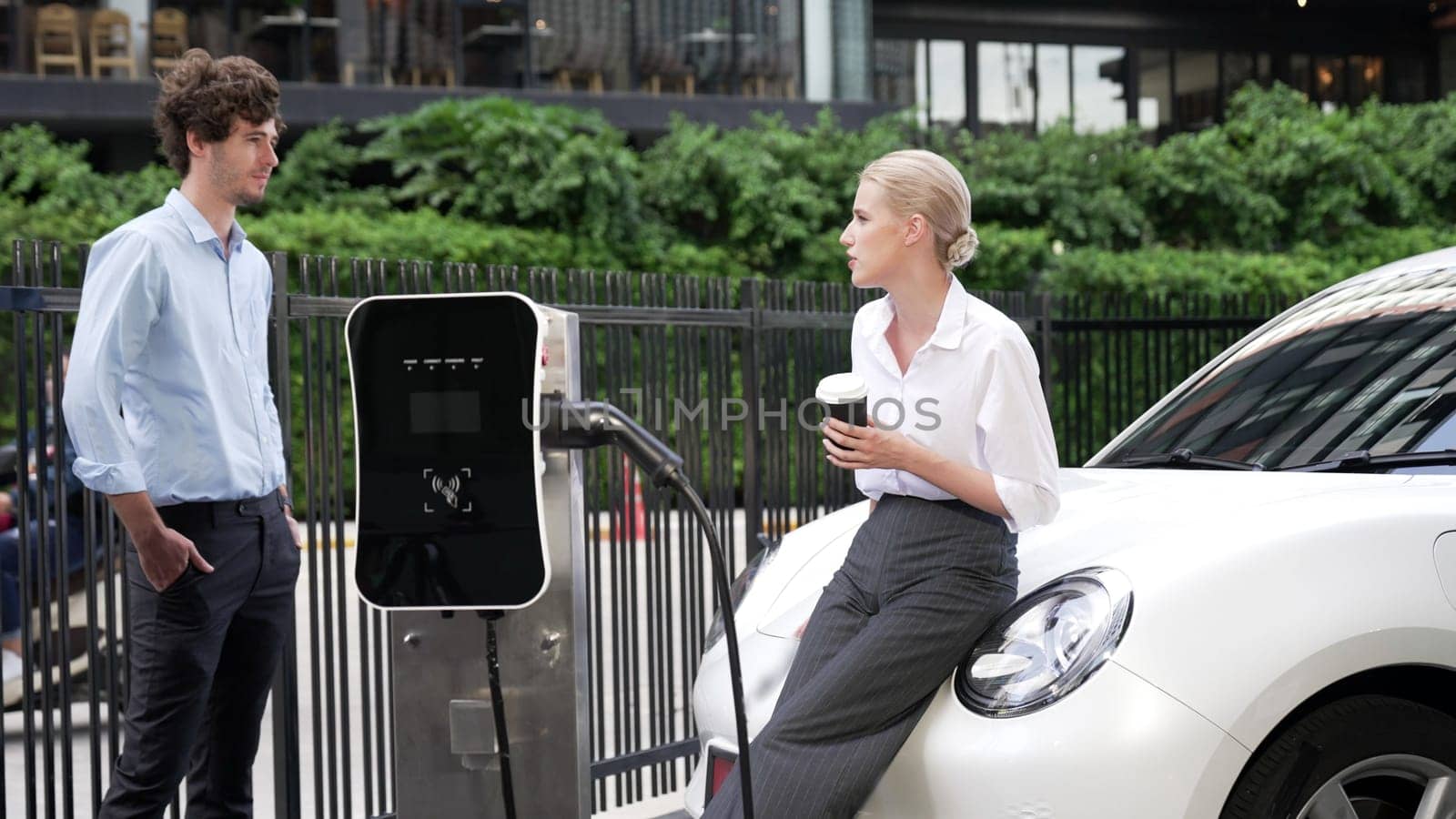 Progressive businessman and businesswoman leaning on electric car connected to charging station before driving around city center. Eco friendly rechargeable car powered by clean energy.
