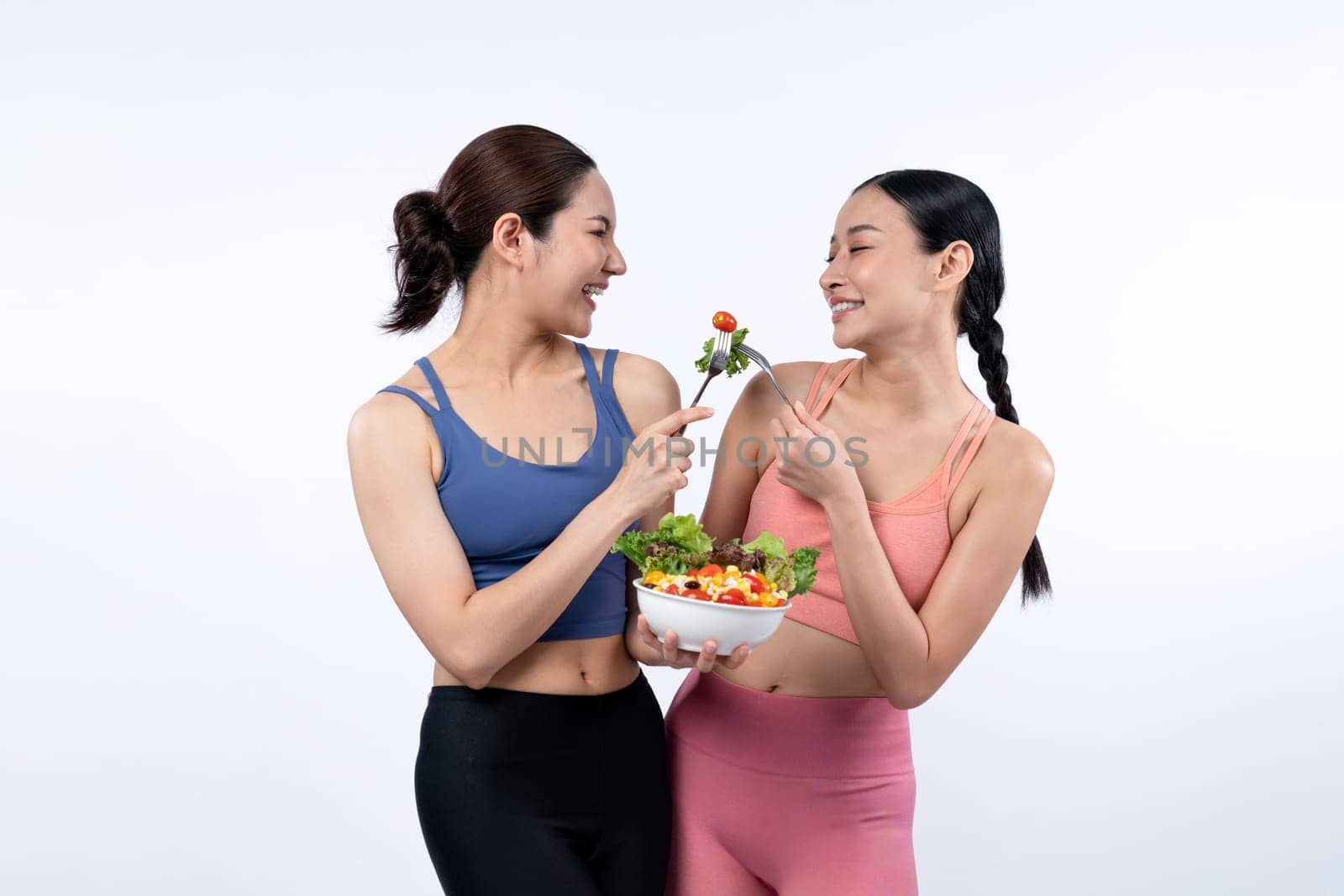 Two young sporty Asian women in sportswear holding salad bowl fill with fruit and vegetable. Natural youthful and fit body lifestyle people with balance nutrition on isolated background. Vigorous