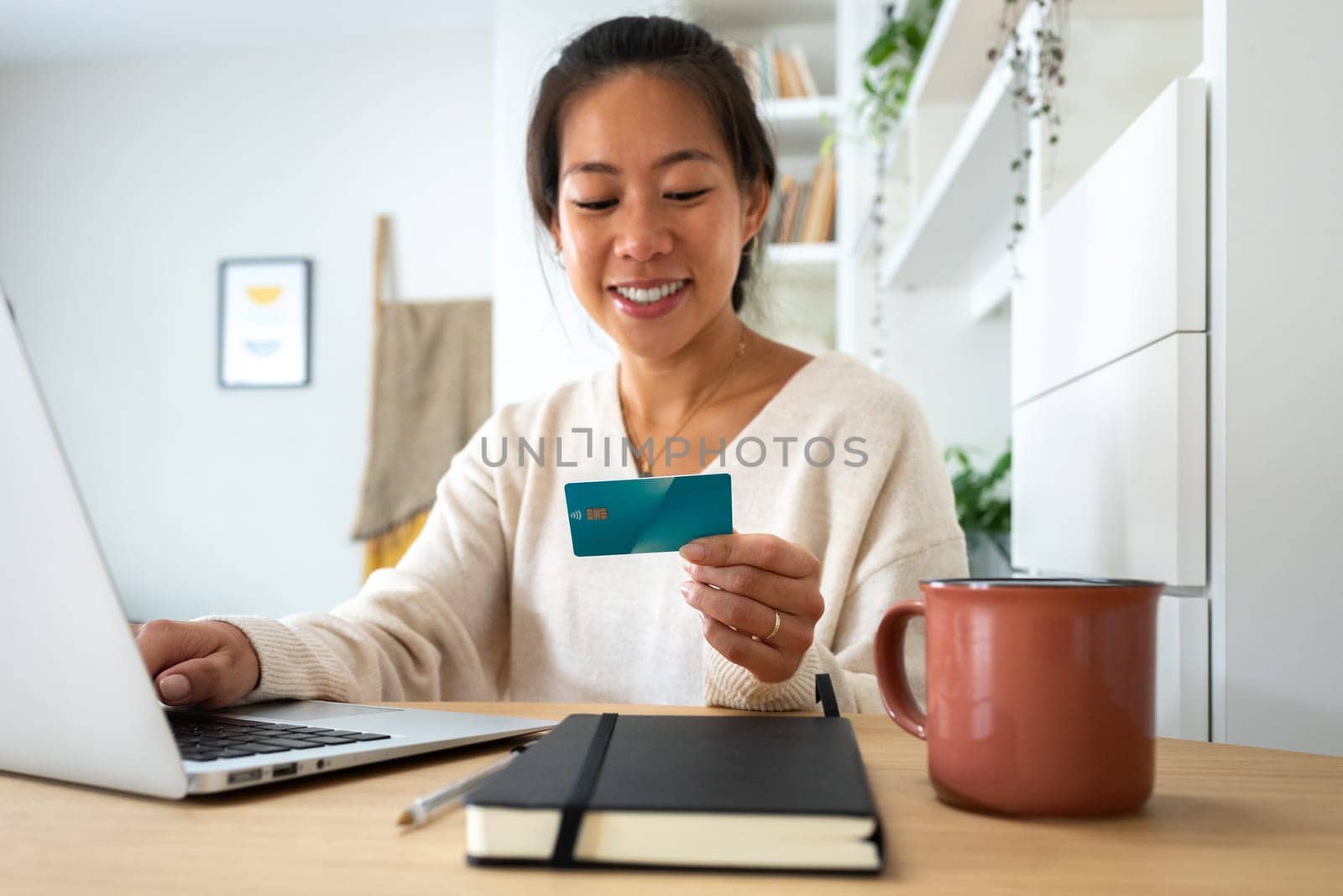 Happy young Asian woman online shopping using laptop and credit card at home office. by Hoverstock