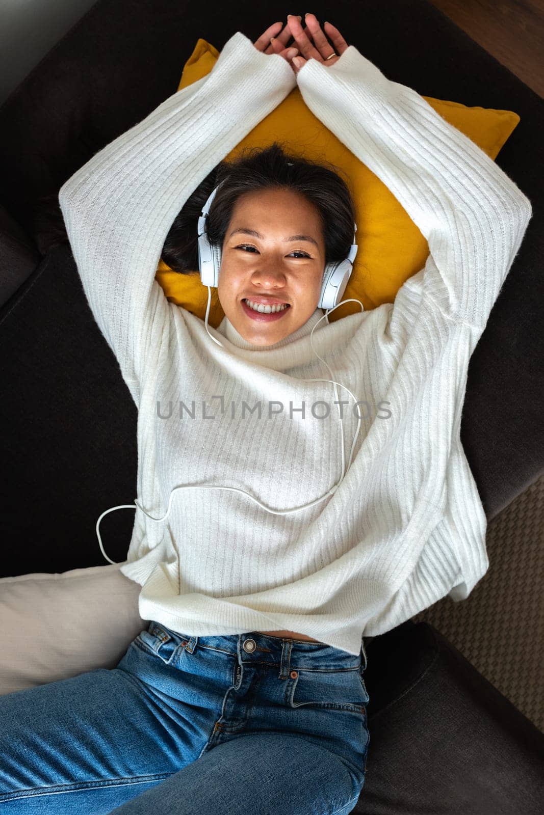 High angle view of young Asian woman lying on the sofa listening to music with headphones looking at camera.Vertical by Hoverstock
