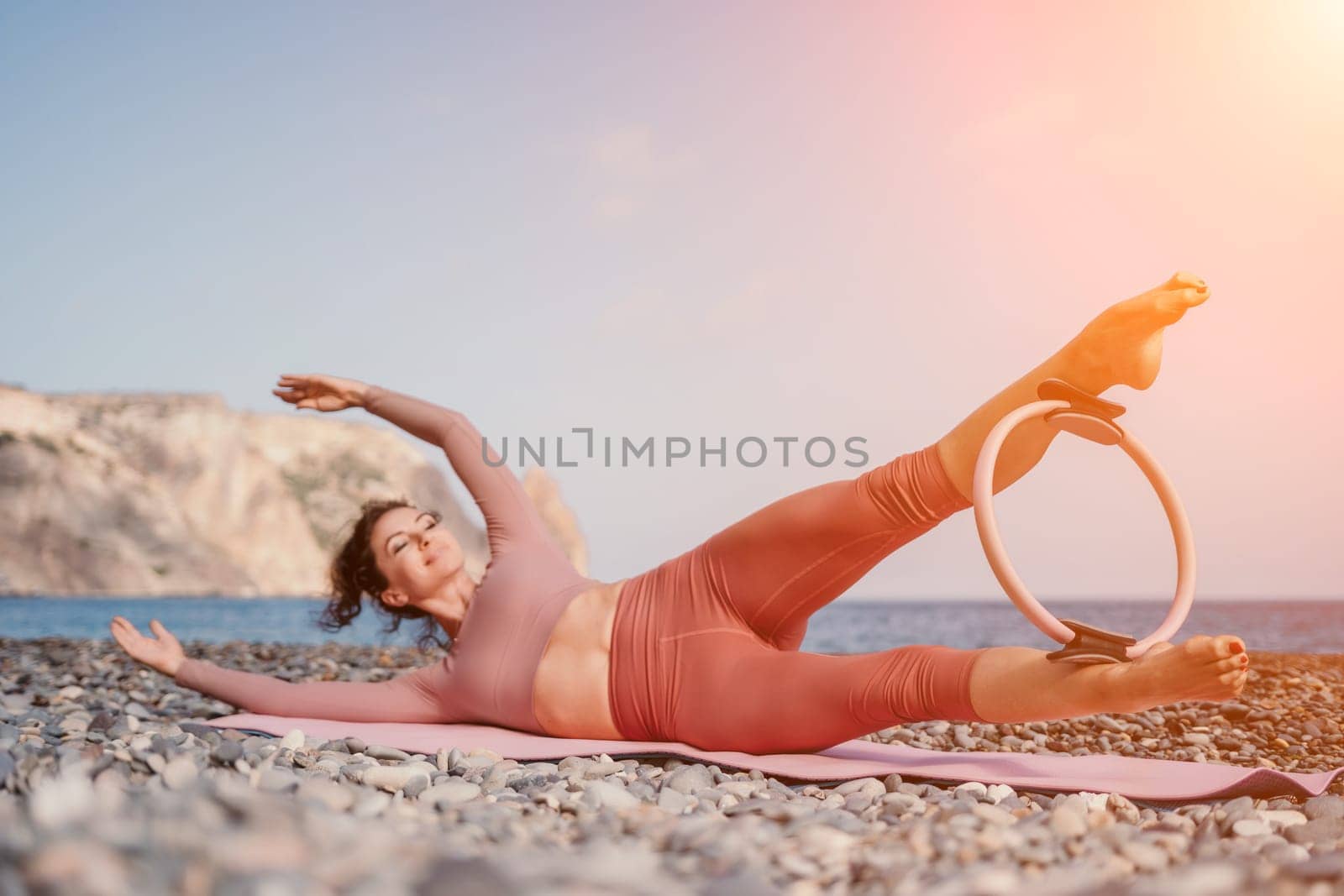 Middle aged well looking woman with black hair doing Pilates with the ring on the yoga mat near the sea on the pebble beach. Female fitness yoga concept. Healthy lifestyle, harmony and meditation.