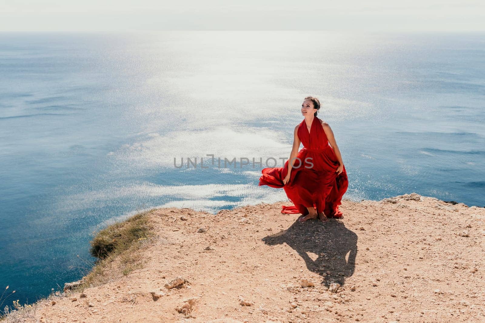 Side view a Young beautiful sensual woman in a red long dress posing on a rock high above the sea during sunrise. Girl on the nature on blue sky background. Fashion photo.