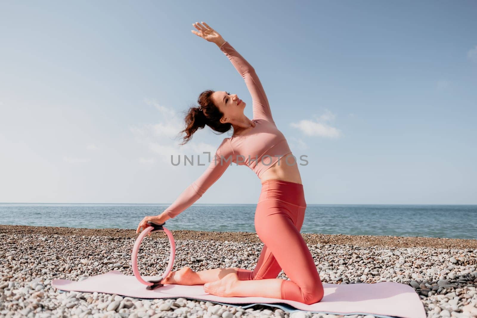 Middle aged well looking woman with black hair doing Pilates with the ring on the yoga mat near the sea on the pebble beach. Female fitness yoga concept. Healthy lifestyle, harmony and meditation.