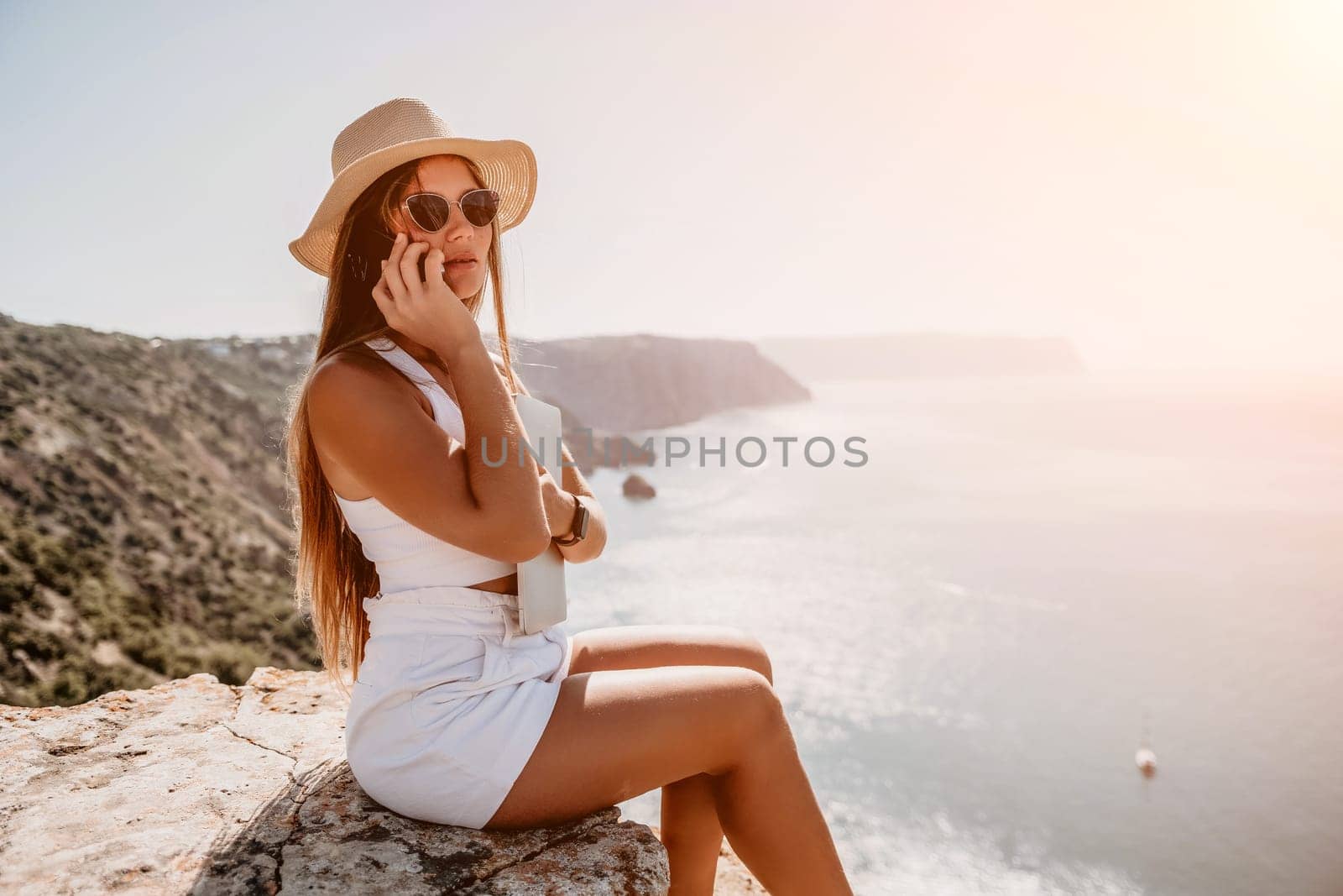 Successful business woman in yellow hat working on laptop by the sea. Pretty lady typing on computer at summer day outdoors. Freelance, travel and holidays concept.