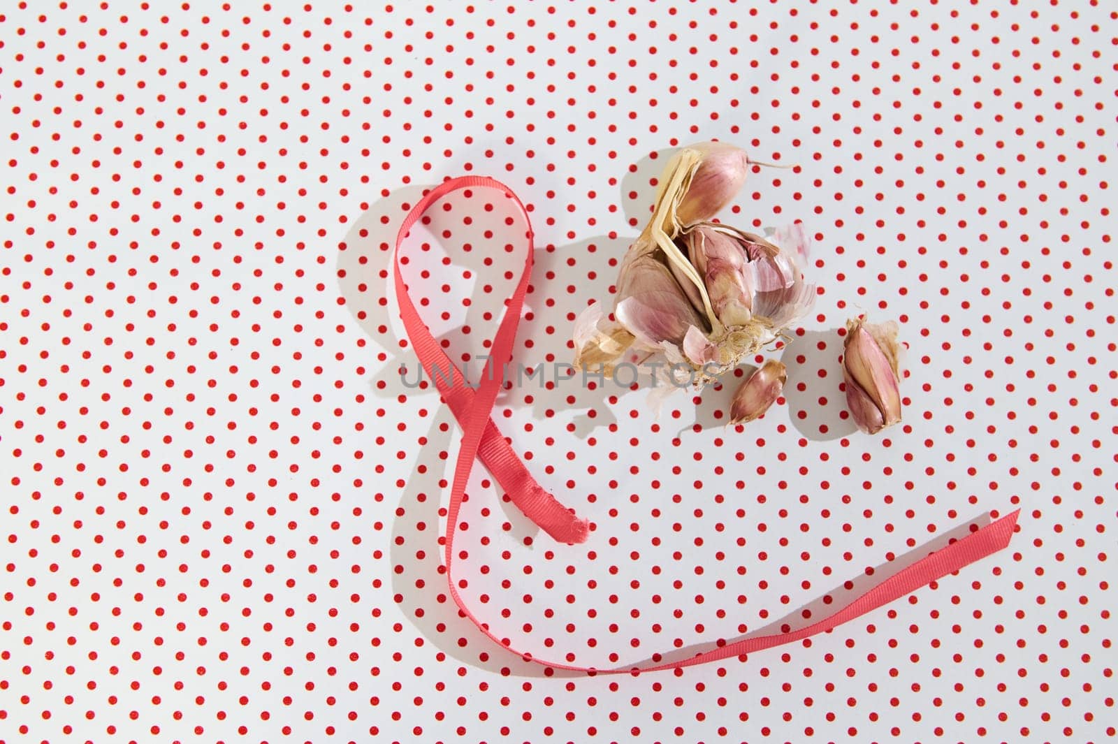 Pink ribbon next to garlic bulbs on a red polka dot backdrop, symbolizing culinary simplicity with visual charm.