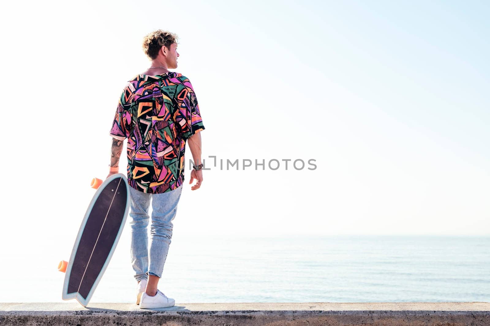 back view of young hipster male skater in trendy summer outfit carrying skateboard while standing on embankment against sea and cloudless sky in summer day