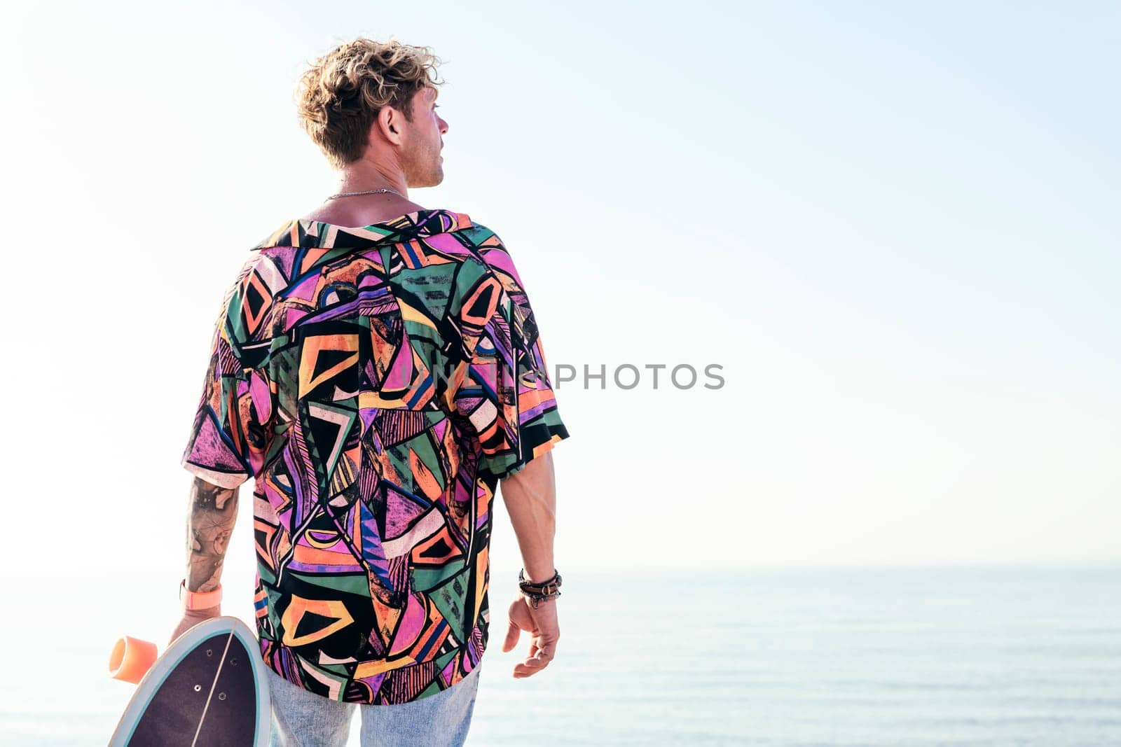 back view of young hipster male skater in trendy summer outfit carrying skateboard while standing on embankment against sea and cloudless sky in summer day
