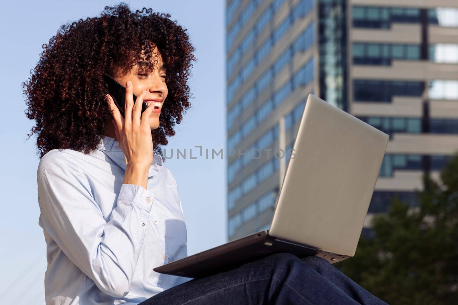 side view of focused happy female entrepreneur sitting at city and working remotely while speaking on smartphone and using laptop