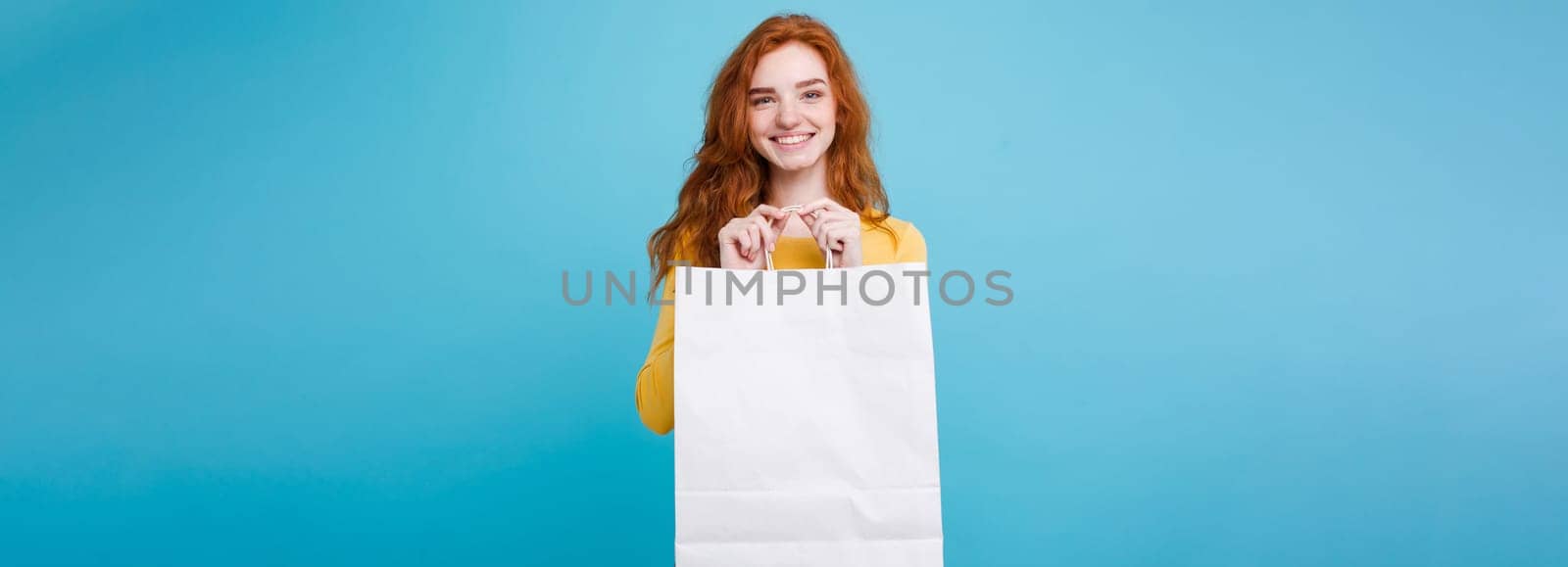 Shopping Concept - Close up Portrait young beautiful attractive redhair girl smiling looking at camera with white shopping bag. Blue Pastel Background. Copy space.