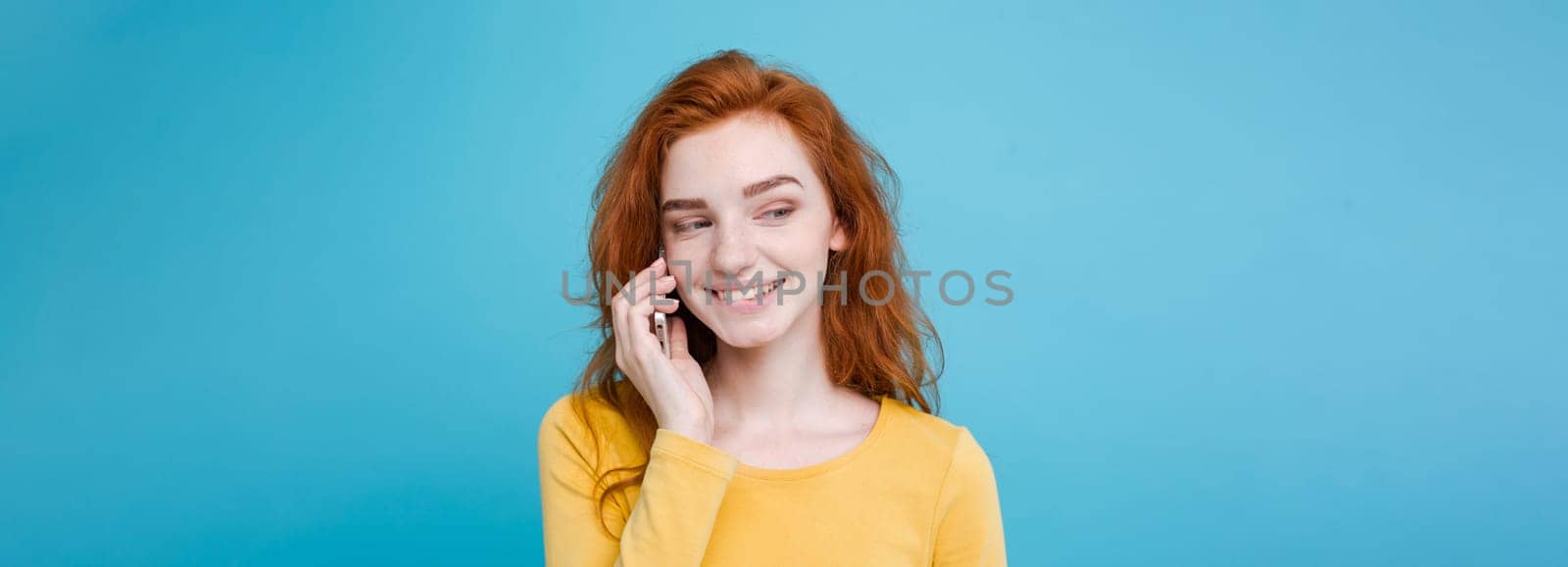 Lifestyle and Technology concept - Portrait of cheerful happy ginger red hair girl with joyful and exciting talking with friend by mobile phone. Isolated on Blue Pastel Background. Copy space.