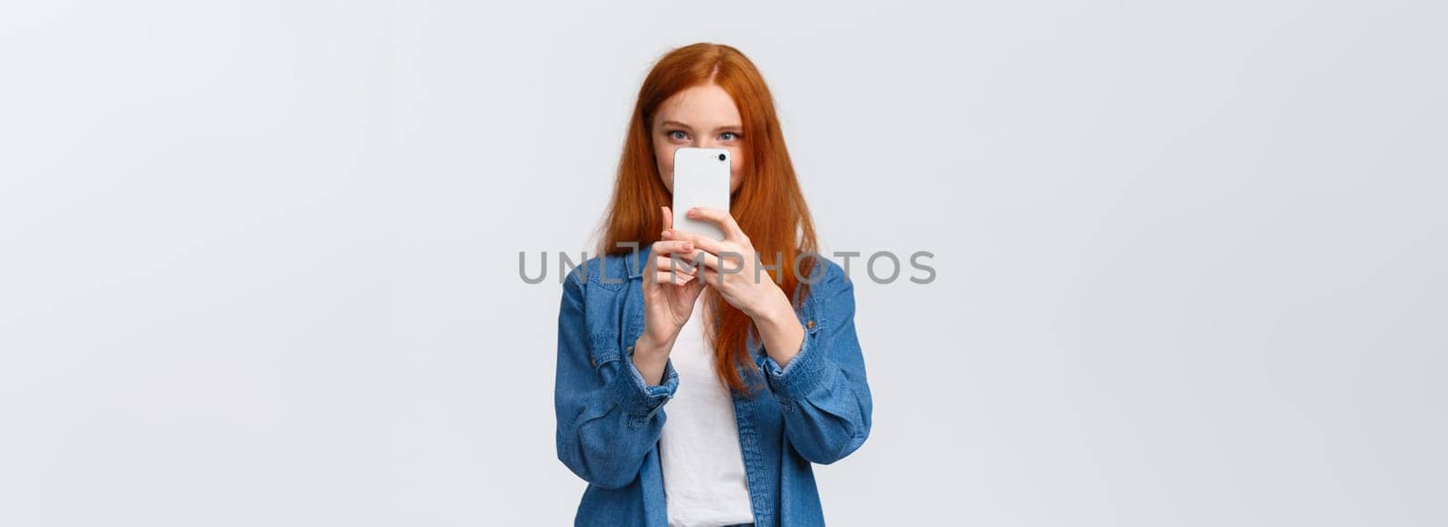 Turning on photo filter. Cheerful and cute female redhead lifestyle blogger, taking picture on smartphone, holding mobile phone near face, taking good shot, searching pretty angle, white background.