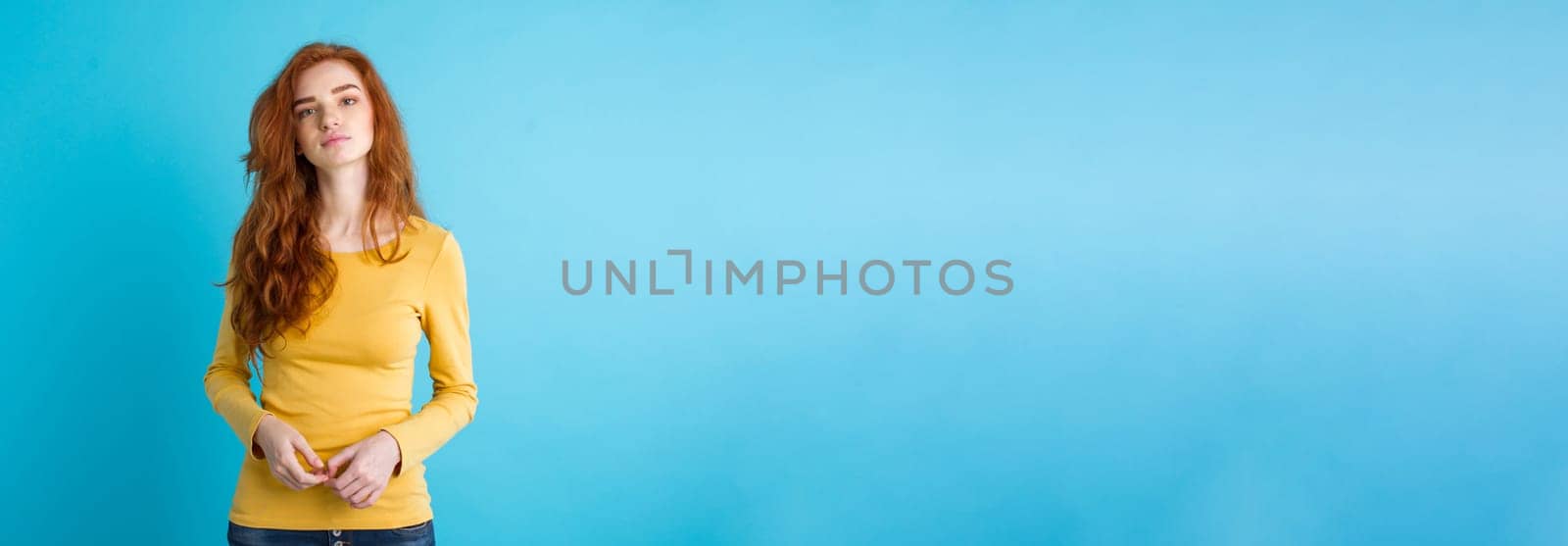 Portrait of young beautiful ginger woman with tender serious face crossing arms looking at camera. Isolated on pastel blue background. Copy space