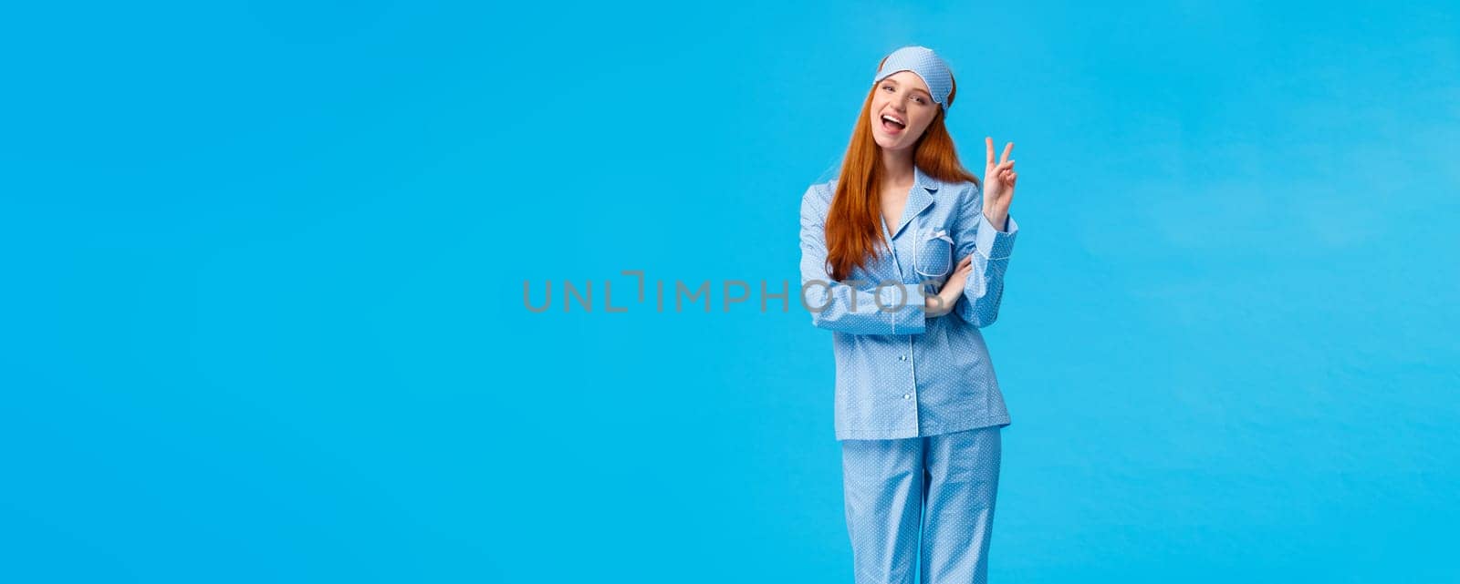 Saying cheeze. Cheerful and cute redhead upbeat girl in glamour pyjama, fancy nightwear having sleepover showing peace sign and smiling excited, having upbeat lucky morning, standing blue background.