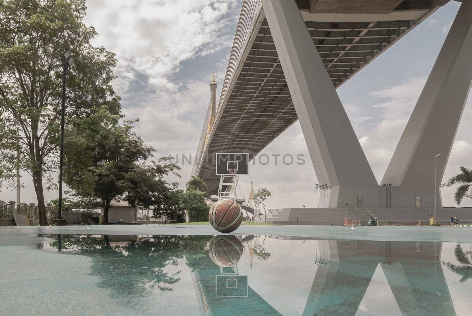 Bangkok, Thailand - 28 Sep, 2024 - Basketball ball on the Old cement court floor with the reflection of them on puddle of rainwater at Outdoor park with Suspension bridge and Sky in the background. Space for text, Selective focus.