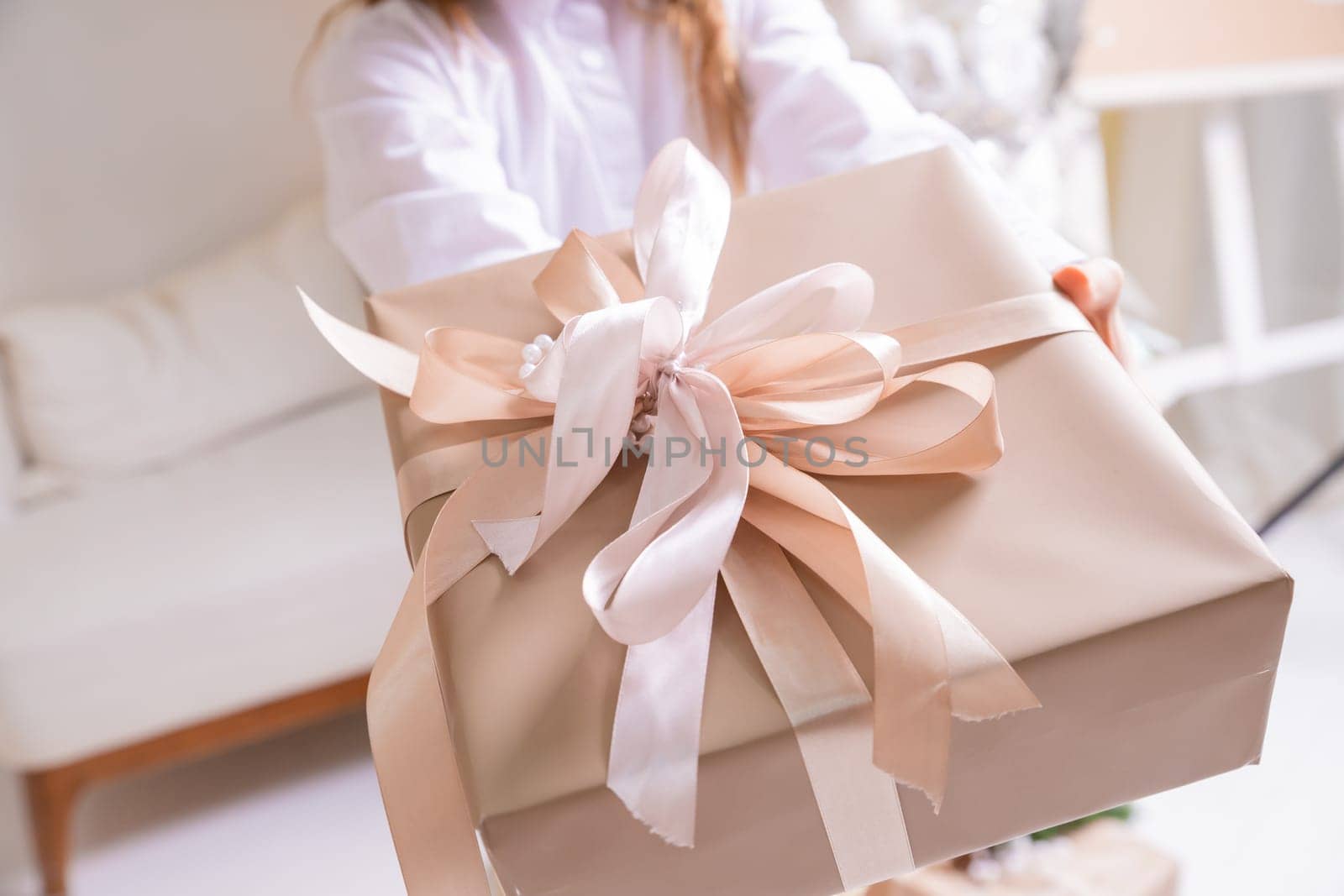 Hands in cozy sweater opening christmas gift with red bow on background of christmas tree with lights.