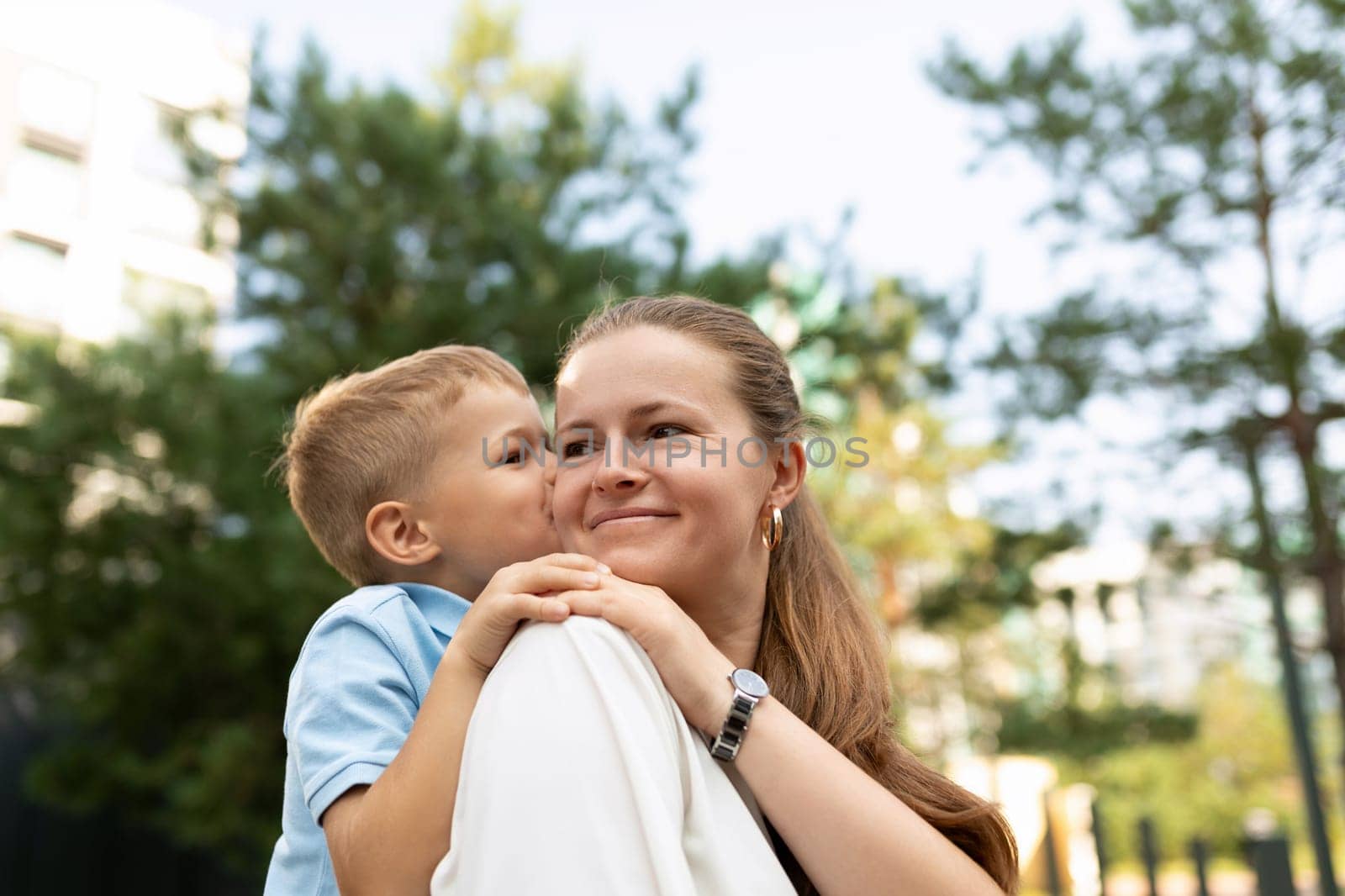A mother smiles warmly as her son cuddles close, sharing a tender moment outdoors. Their bond shines brightly under the sun in a vibrant park setting.