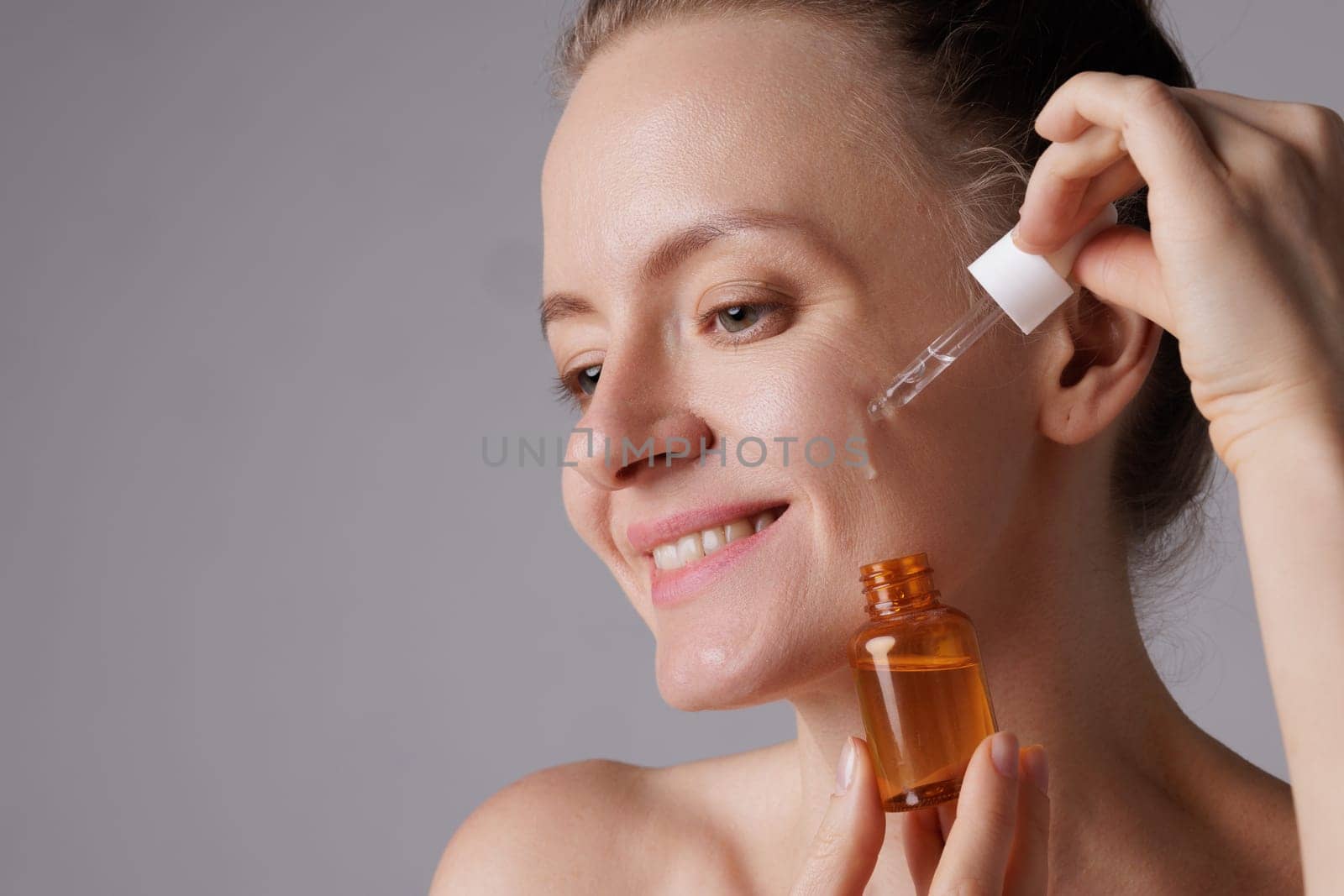 Happy smiling woman applying serum on her face, close eyes with pleasure. Redhead girl holding dropper with skin care product for healthy and glowing skin tone, blue background
