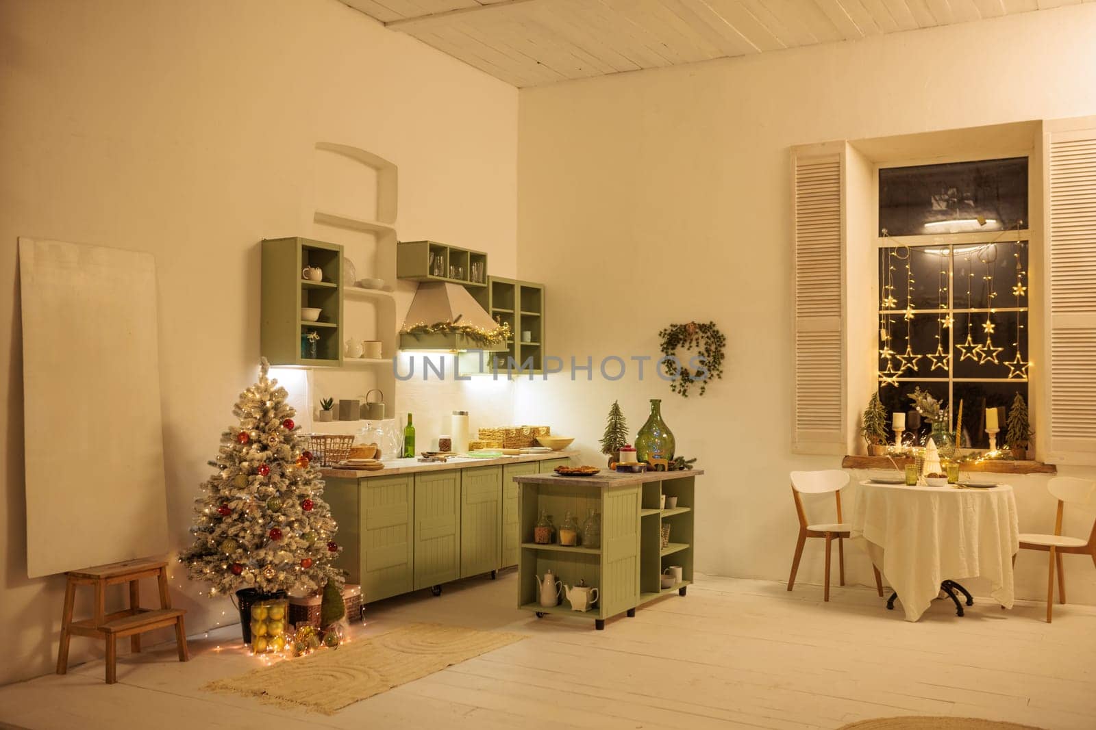 A cozy, bright kitchen decorated for Christmas with green cabinets, a small Christmas tree with decorations, and a festive table with snacks.