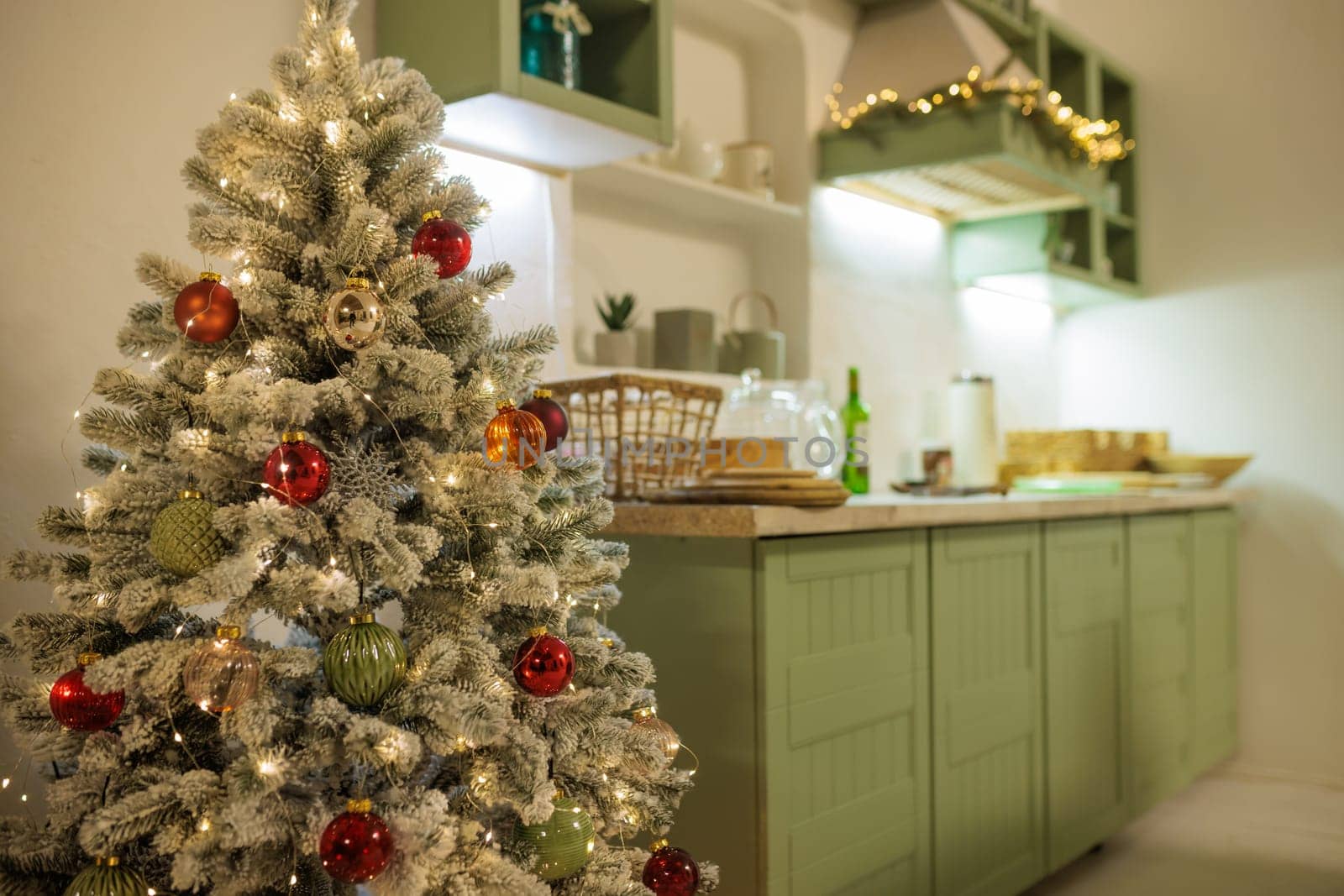 A cozy, bright kitchen decorated for Christmas with green cabinets, a small Christmas tree with decorations, and a festive table with snacks.