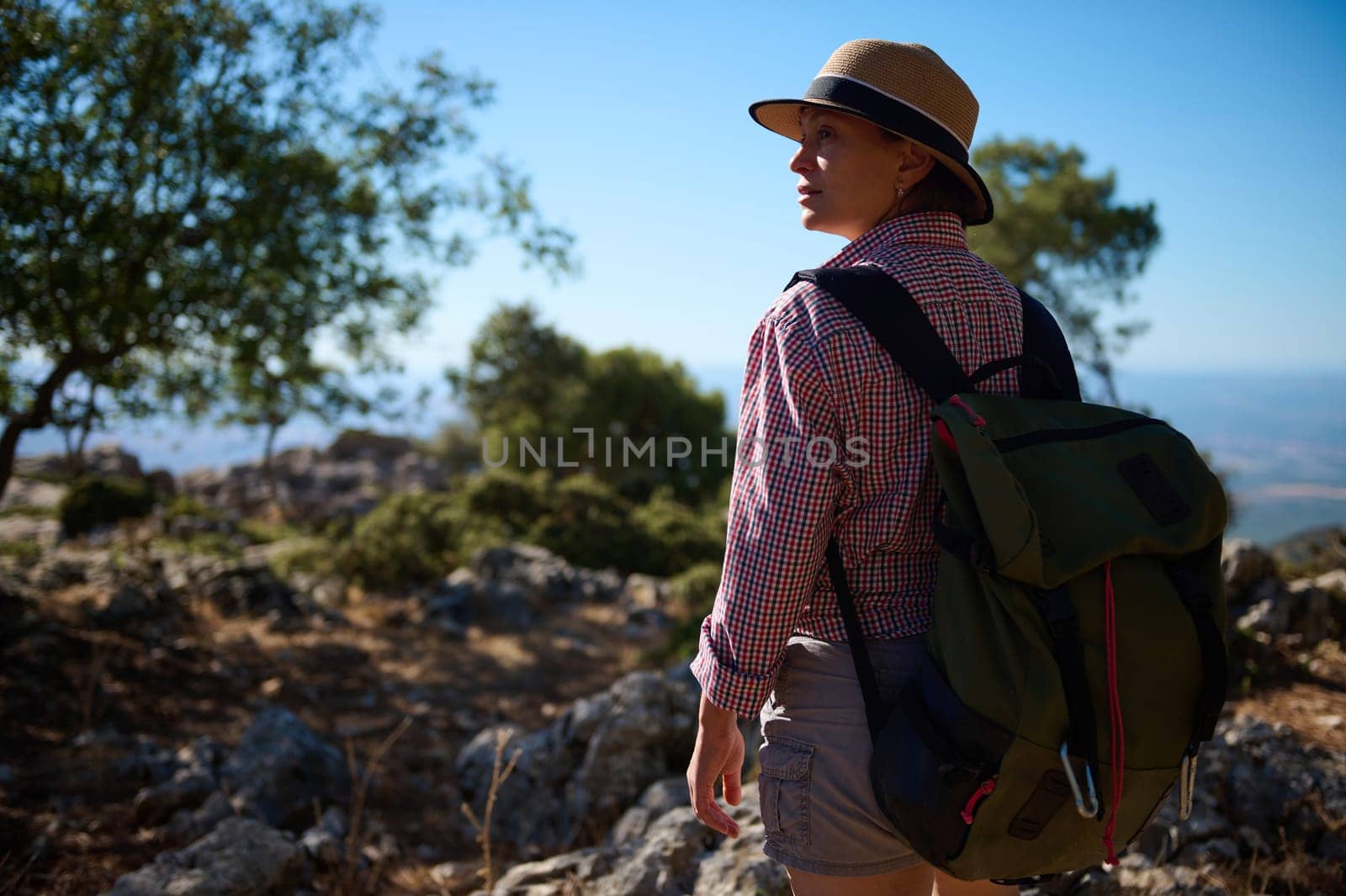 A woman in a hat and backpack explores a rocky terrain during a hike on a sunny day. The vibrant landscape and clear blue sky create an atmosphere of adventure and discovery.
