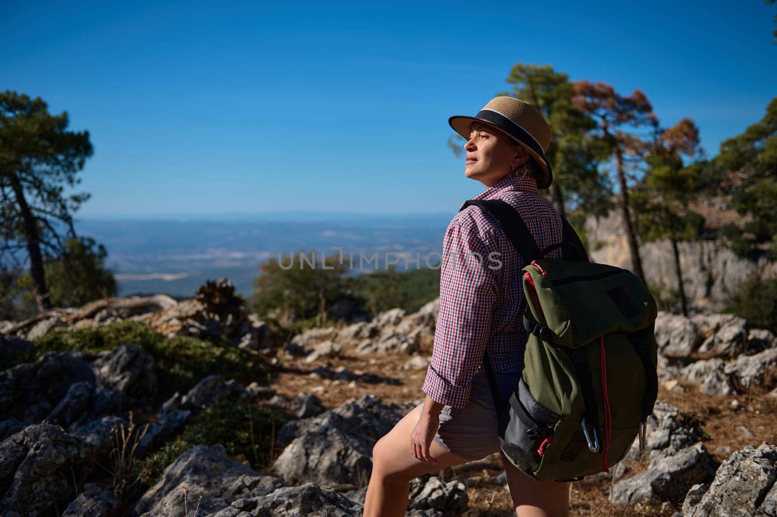 A woman enjoys a hike through rocky terrain, carrying a backpack and wearing a sun hat. The clear blue sky and expansive view highlight the adventurous spirit of outdoor exploration.