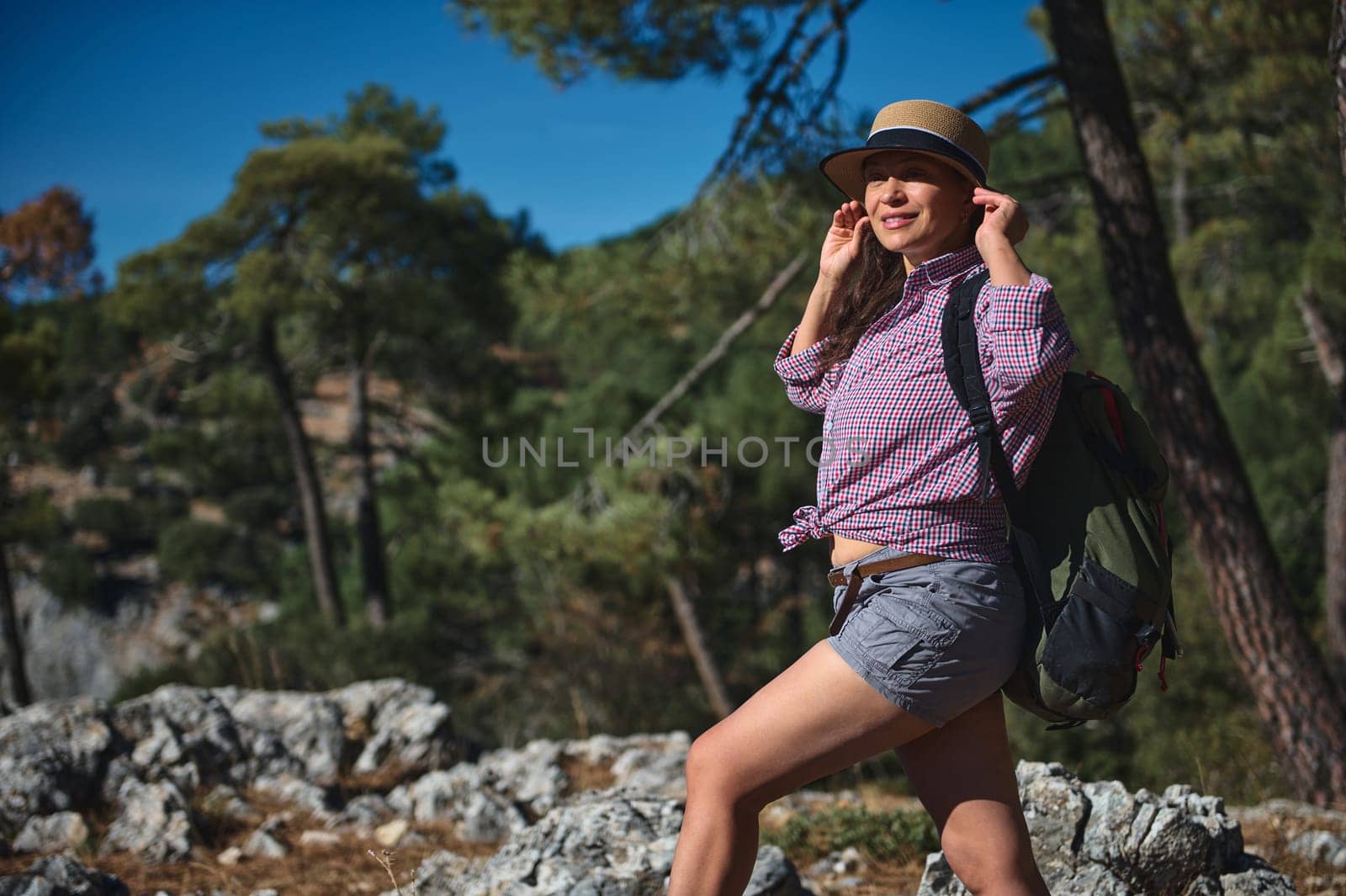 A woman in a hat and casual attire enjoys a hike in a sunlit forest, capturing the essence of adventure and nature. The lush greenery and rugged terrain highlight her journey.
