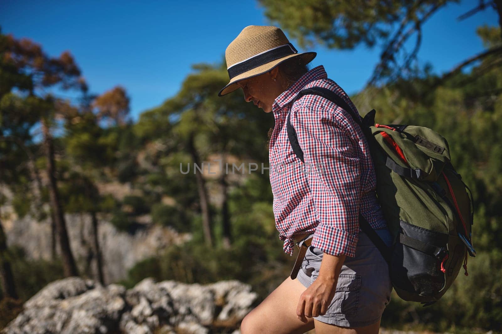 A woman wearing a hat and carrying a backpack hikes through a sunlit forest. The outdoor setting and casual attire suggest a relaxing and adventurous experience in nature.