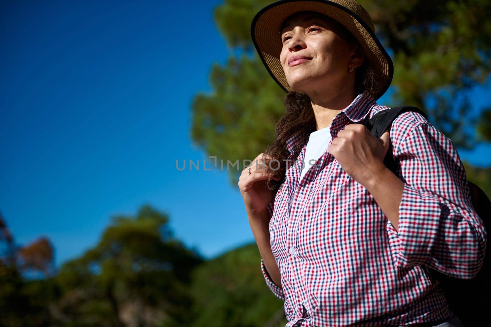 A confident woman enjoys hiking in a sunny outdoor landscape. She wears a hat and backpack, looking content and adventurous. The blue sky adds to the tranquil setting.