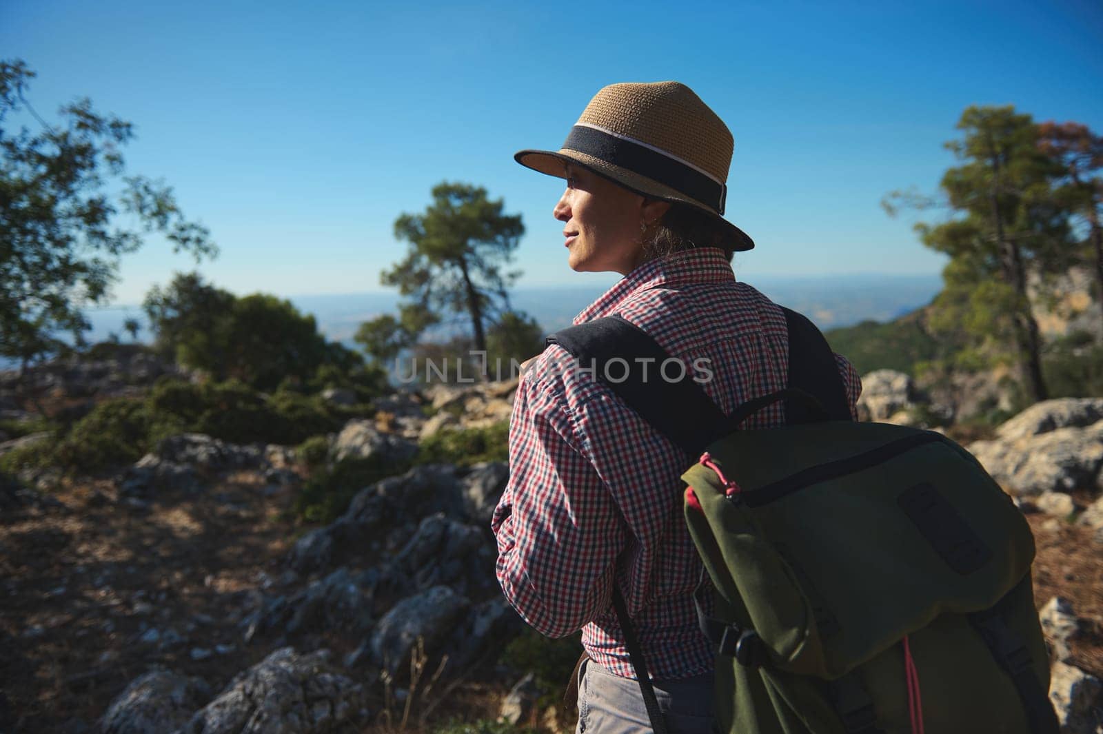 A woman in a checkered shirt and hat enjoys a sunny mountain hike. She carries a backpack, embracing the tranquility of the natural landscape surrounding her.