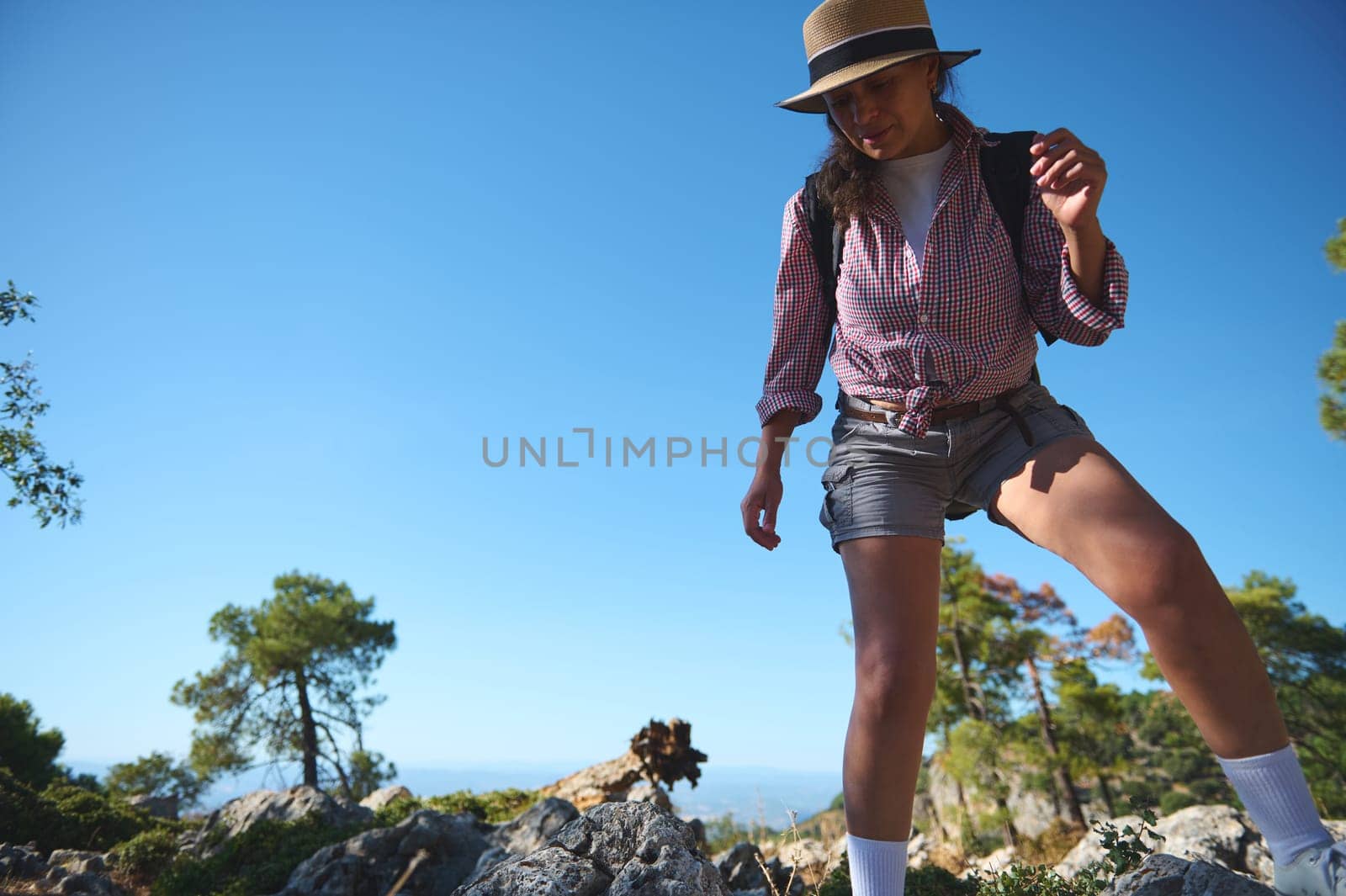 A young woman wearing a hat and carrying a backpack hikes through a rocky landscape. She is joyfully exploring a mountainous area on a sunny day, capturing the essence of adventure.