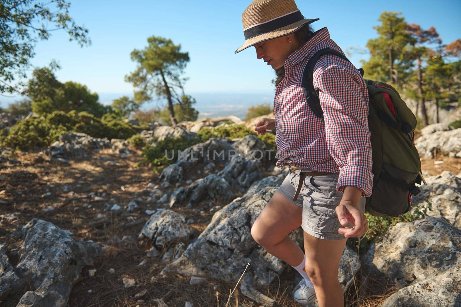 A woman in a hat and checkered shirt hikes through rocky terrain with trees in the background. She carries a backpack, symbolizing adventure and exploration in a natural setting.