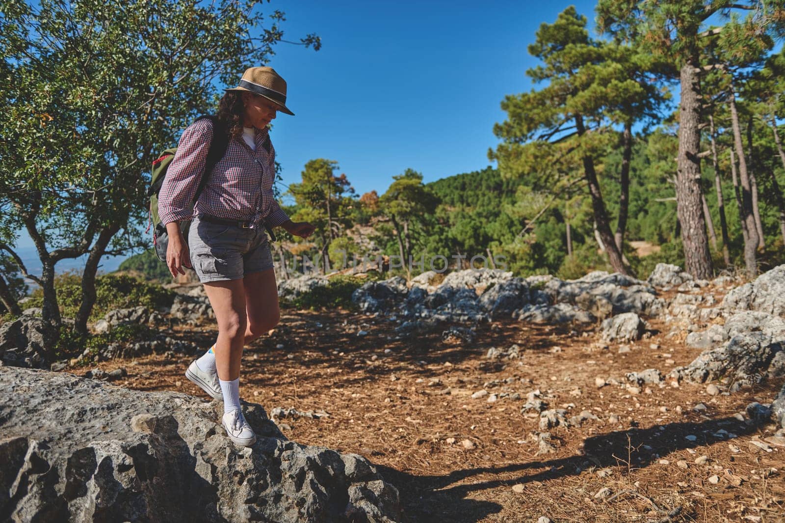 A woman wearing a hat and backpack hikes along a rocky path through a scenic forest. The sunny day highlights the lush greenery and tranquil setting, perfect for nature exploration.