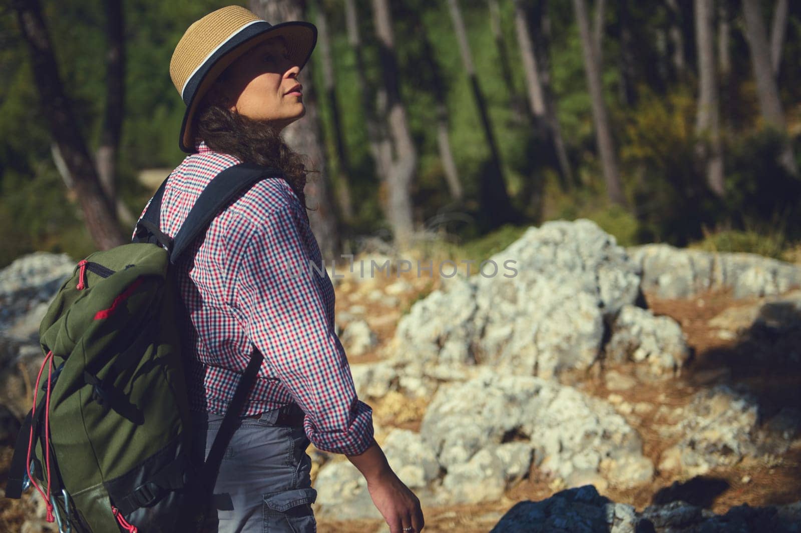 A young woman enjoys a peaceful hike through a sunlit forest, carrying a backpack and wearing a straw hat. She admires the natural scenery and feels a deep connection with nature.