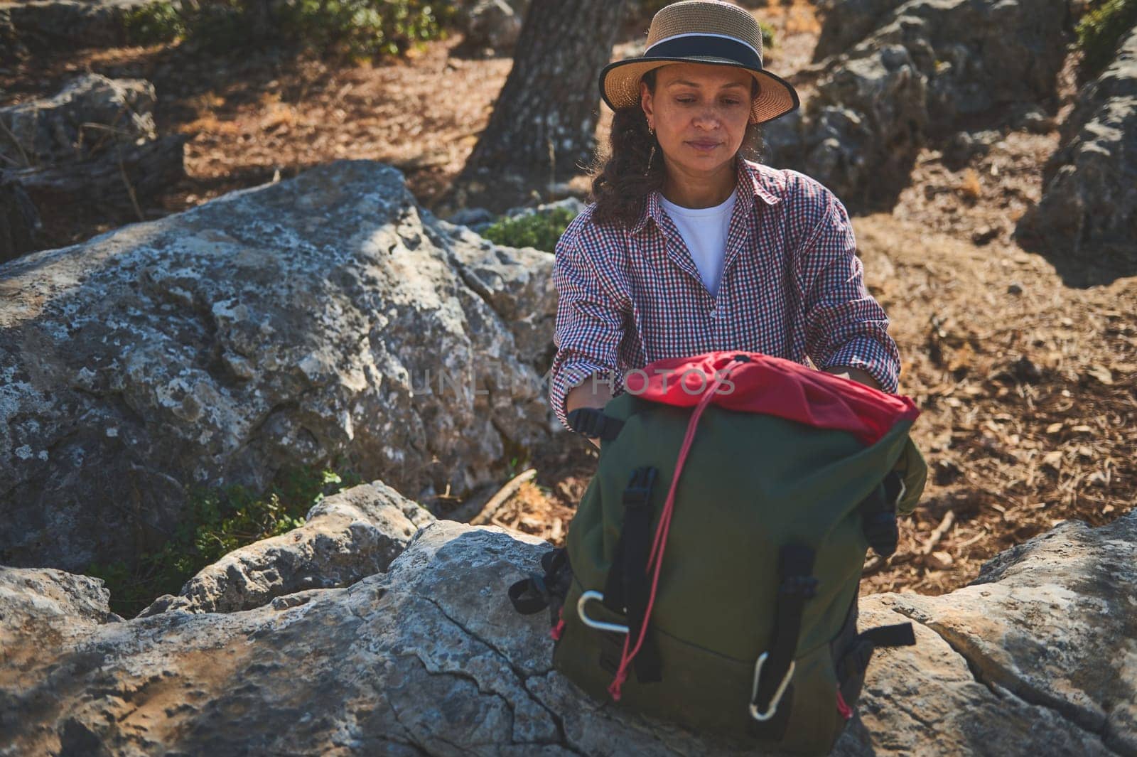 A woman in a hat and checked shirt prepares her green backpack on rocky terrain. Surrounded by nature, she embodies the spirit of adventure and exploration, enjoying a peaceful hiking experience.