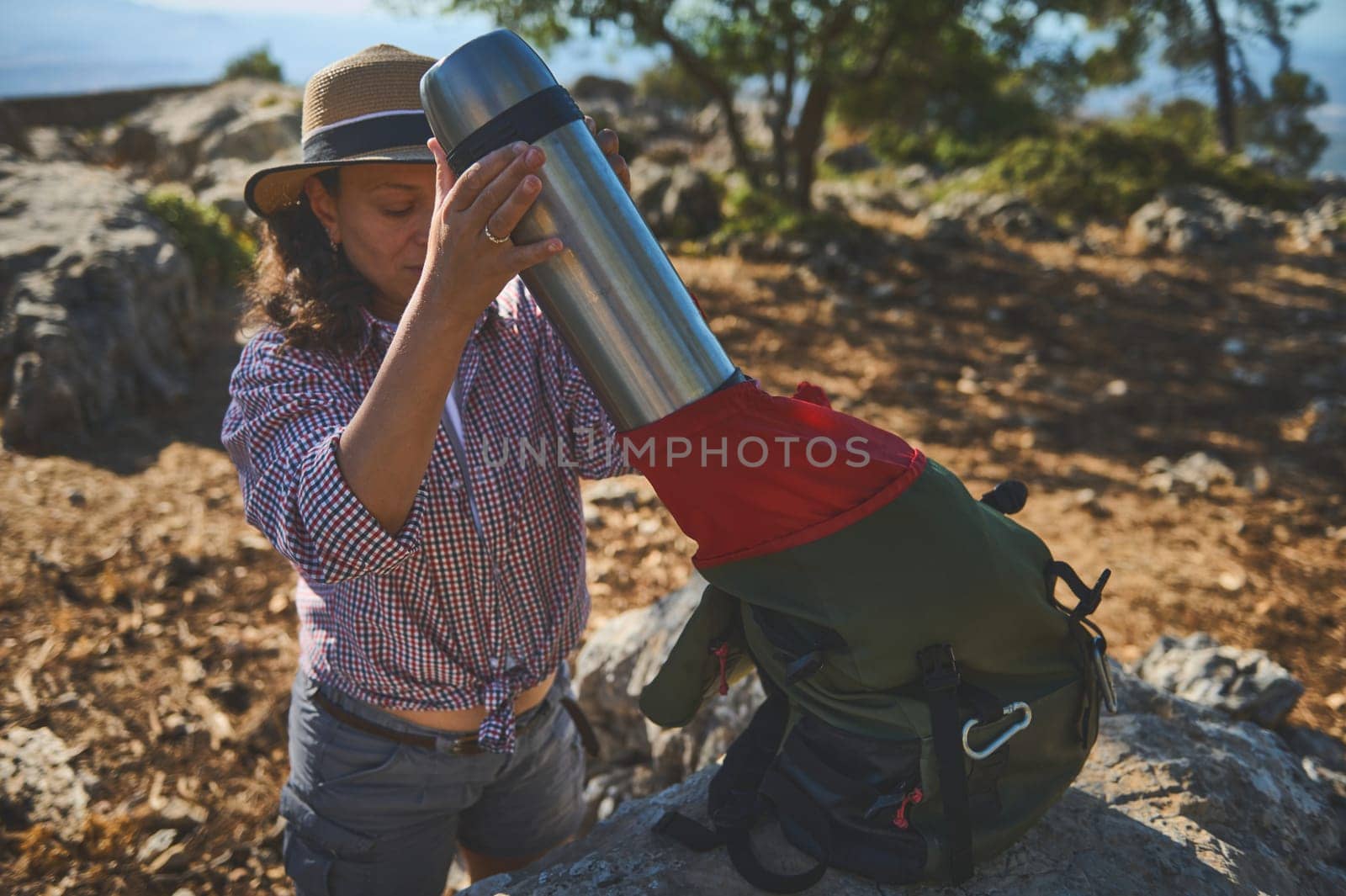 A woman equips herself for a hiking adventure, packing a large thermos into her backpack, in a rocky outdoor setting under the sun.