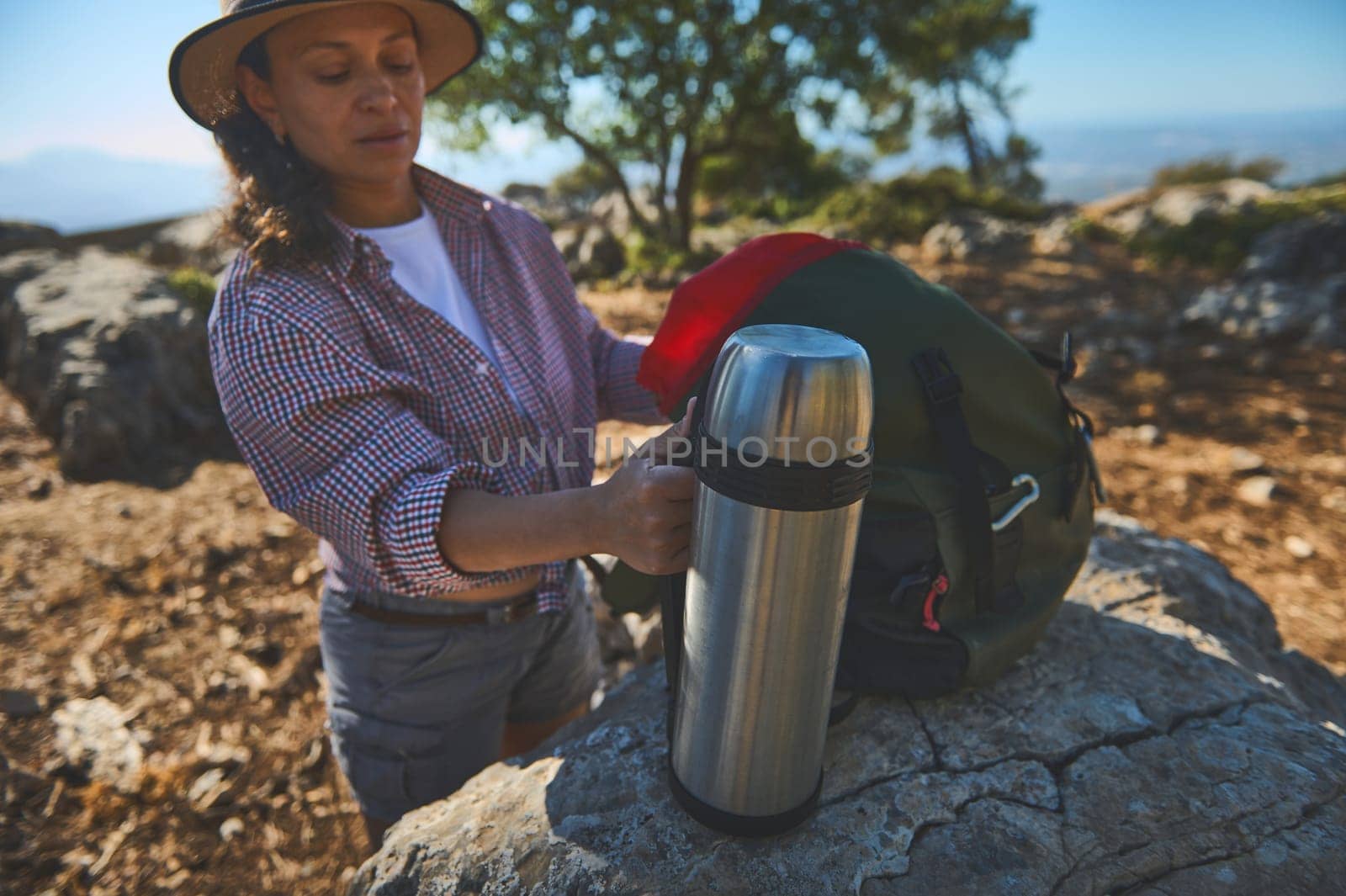 A woman takes a break during a scenic mountain hike, holding a thermos beside her backpack. The peaceful outdoor setting highlights a love for nature and adventure.