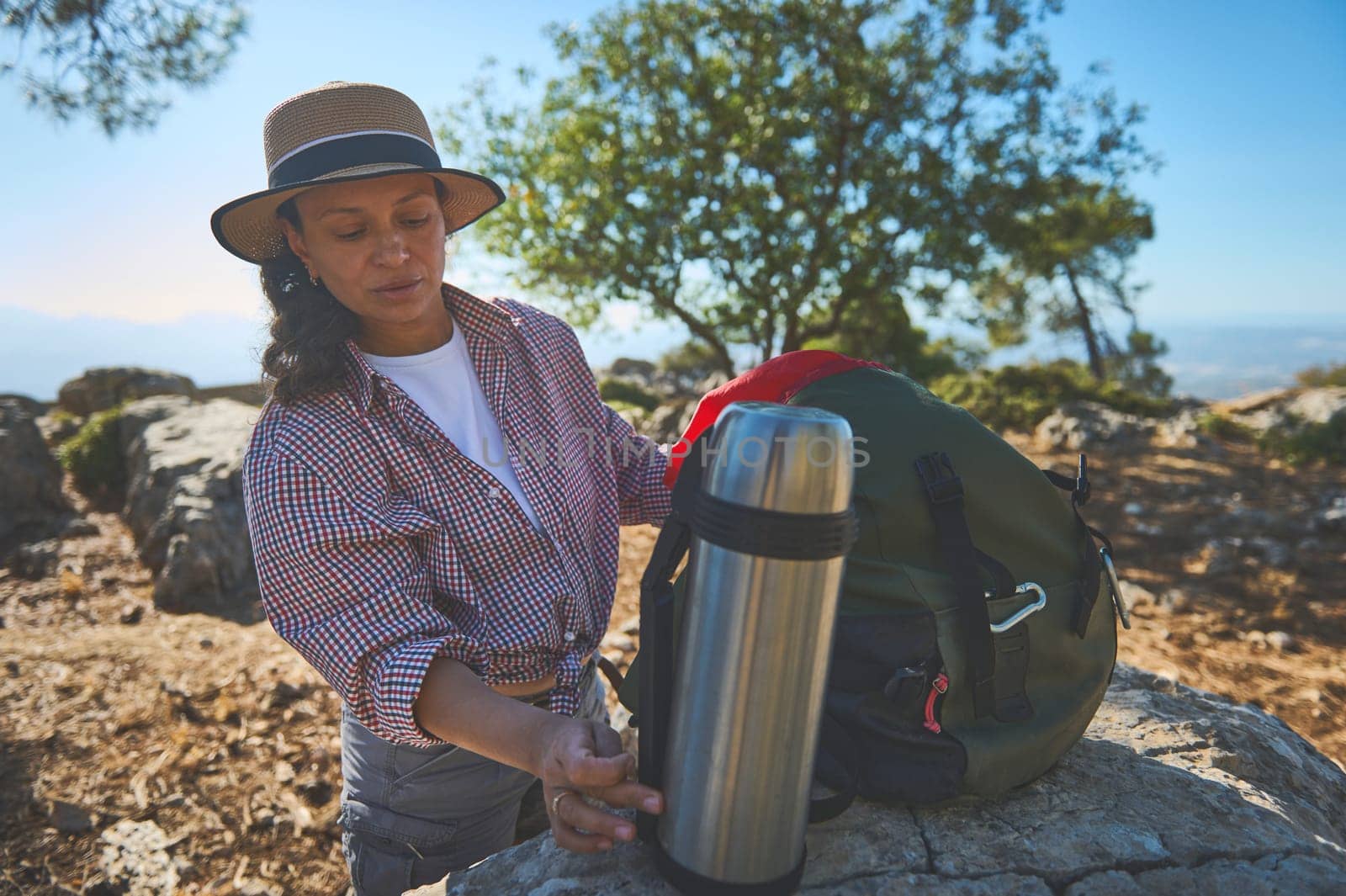 A woman dressed in a hat and checkered shirt prepares for a hike, standing beside a backpack with a thermos. The scenic mountain view and sunny weather enhance the outdoor adventure.