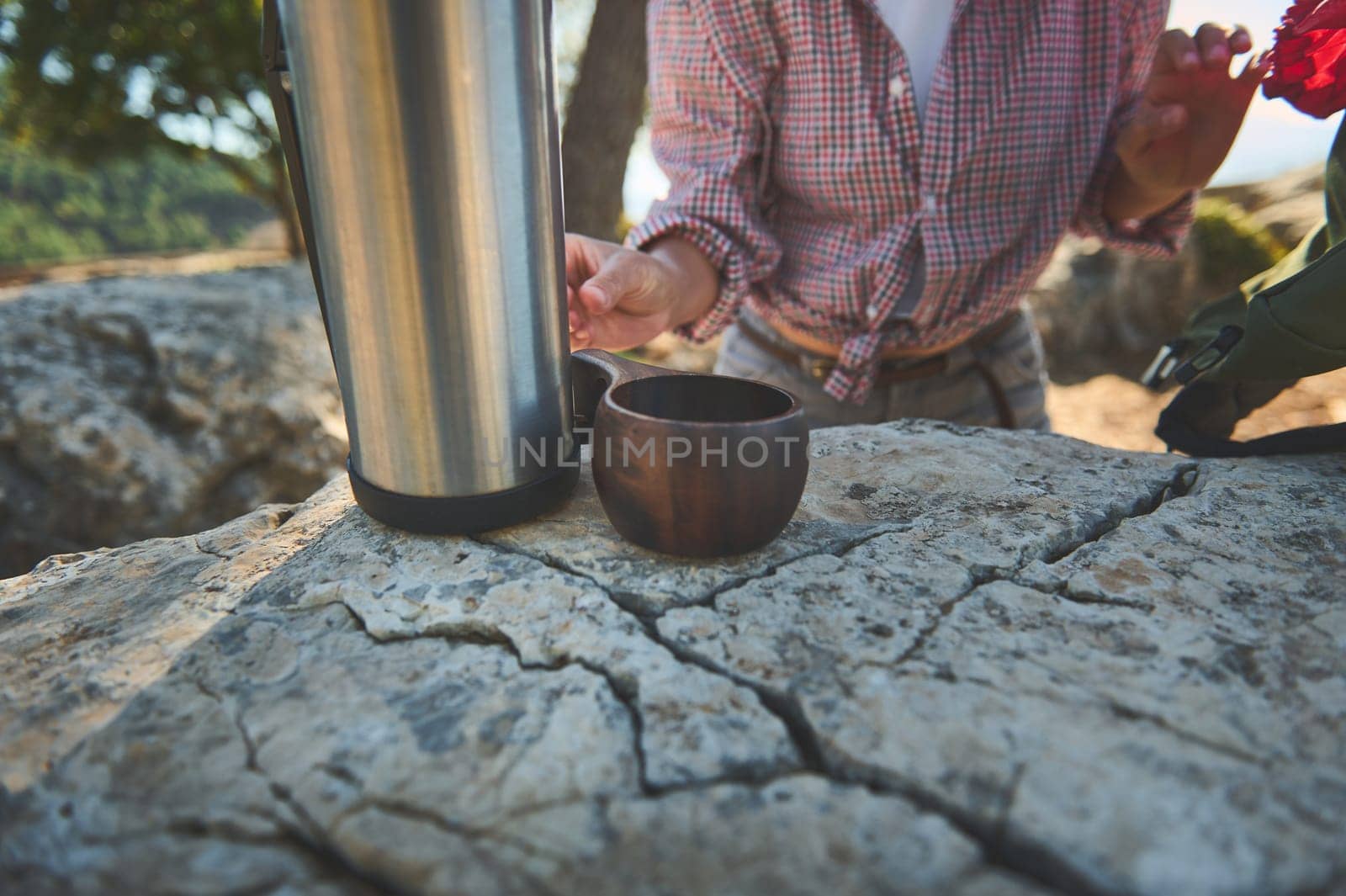 A person in a plaid shirt prepares a hot drink from a thermos on a rocky outdoor surface, embracing nature.