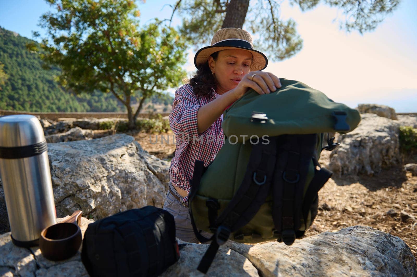 A woman prepares a backpack among rocks and trees, ready for a hiking adventure. Sunlight filters through the greenery, creating a serene atmosphere. An ideal setup for an outdoor escape.
