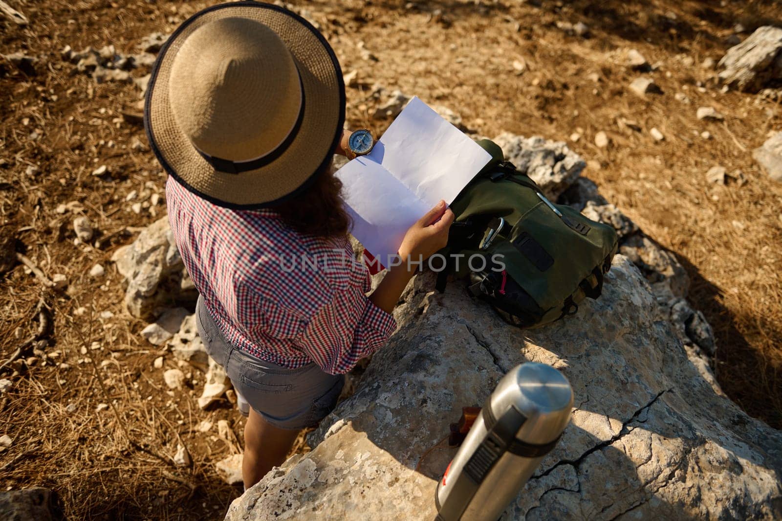 A woman in a hat and checked shirt reading a map while sitting on a rocky terrain. She is surrounded by a travel backpack and a thermos, enjoying the sunny outdoors.