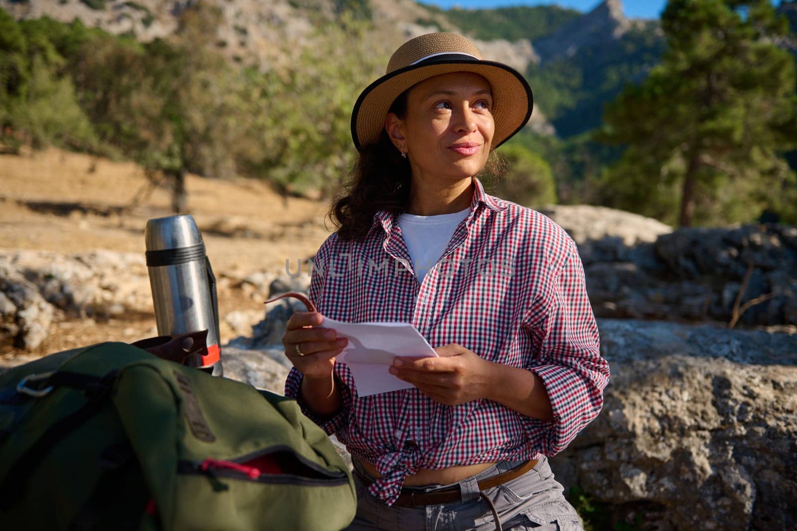 A woman wearing a straw hat and plaid shirt sits on a rock holding a letter, enjoying a tranquil moment in nature. A thermos and backpack are nearby, evoking a sense of adventure.