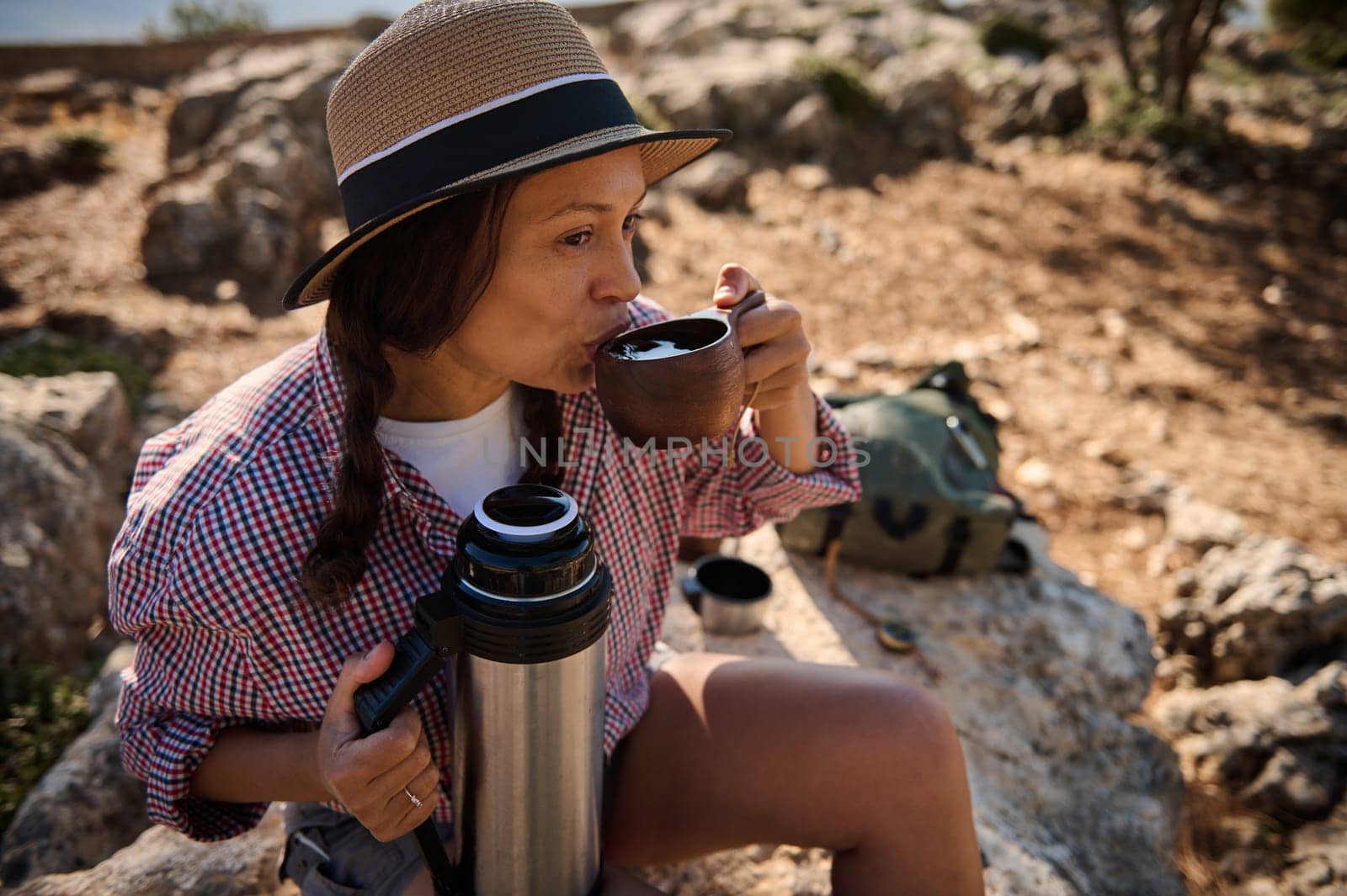 A woman in a straw hat sips a hot drink while seated on a rock. Surrounded by nature, she holds a thermos, capturing a peaceful outdoor moment.