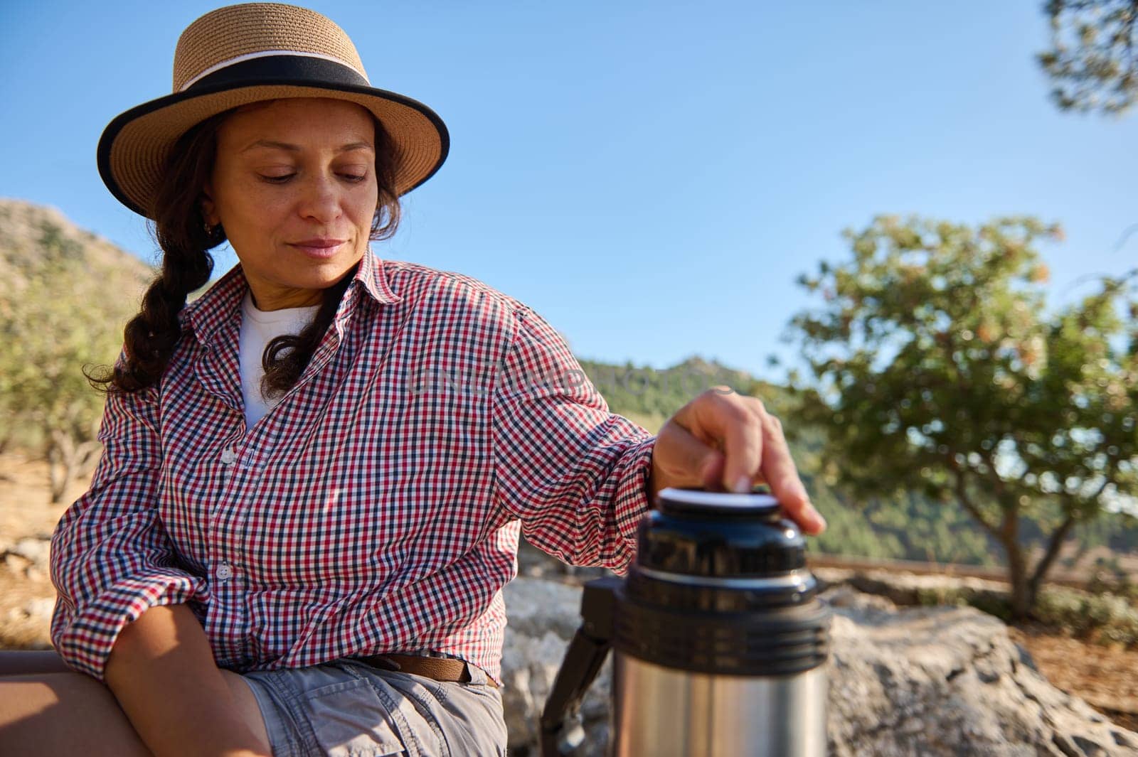 A woman sitting outdoors, wearing a hat and checkered shirt, enjoys a calm moment with a thermos. The natural setting, warm sunlight, and relaxed posture convey tranquility and leisure.