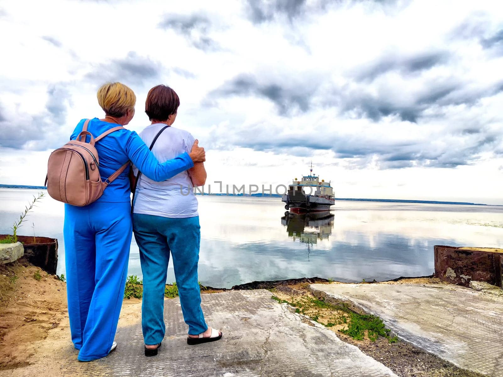 Two women stand together, watching a boat reflected on the water while enjoying a cloudy day.