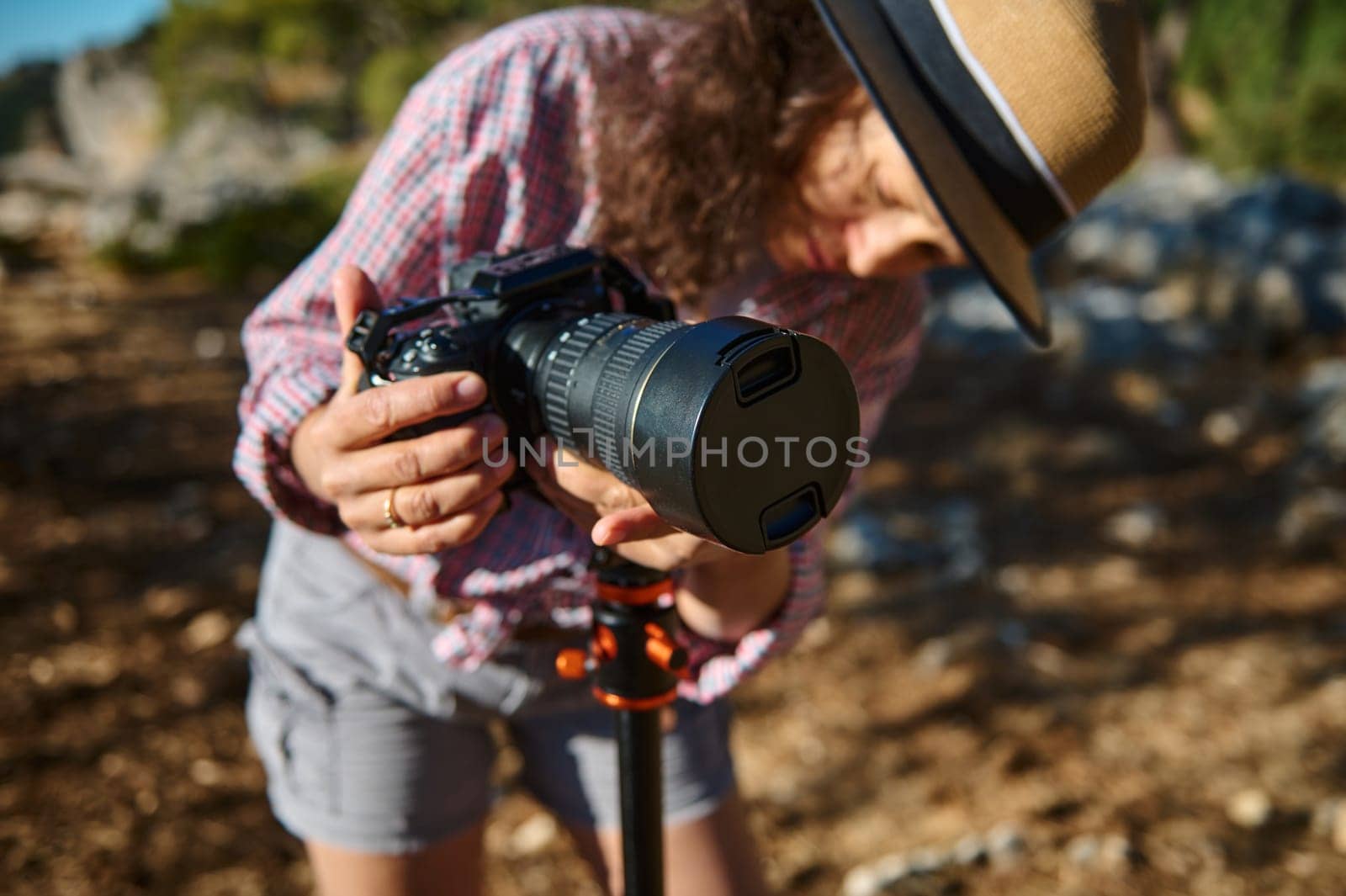 Close-up of a woman in a hat adjusting a camera on a tripod in a scenic outdoor environment, capturing the beauty of nature.