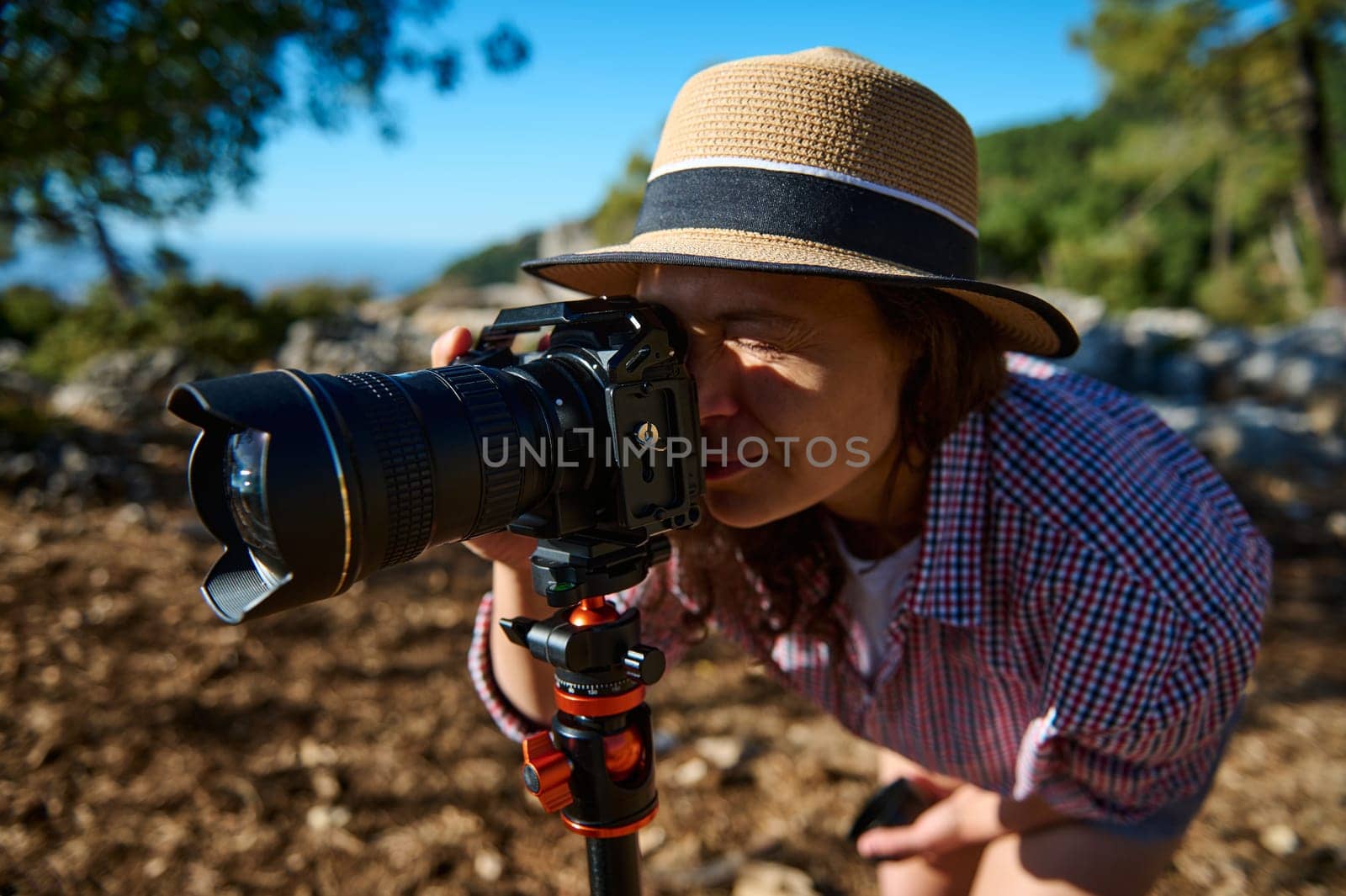 A woman wearing a straw hat and checkered shirt uses a camera mounted on a tripod to photograph nature in a sunny outdoor setting. The background is a scenic natural landscape.